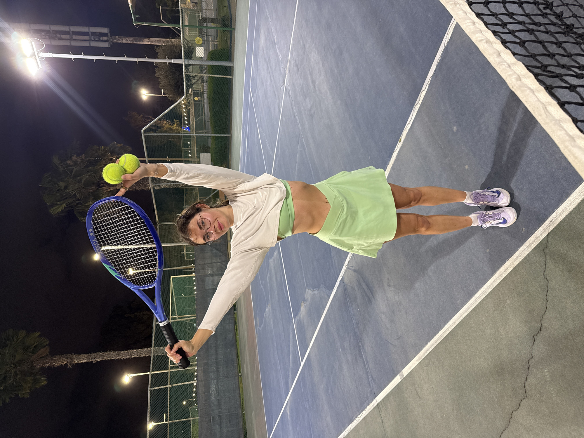 A young woman stands on an outdoor tennis court at night, holding a tennis racquet in her right hand and two tennis balls in her left. She wears a light-colored long-sleeved shirt, a green sports bra, and a matching green tennis skirt. Her footwear consists of athletic shoes. The tennis court has a blue and gray surface with white lines and a black net visible on the right side. In the background, there are tall palm trees and street lights illuminating the area, suggesting a city environment. Given the prompt, the location is assumed to be in Tel-Aviv, Israel.