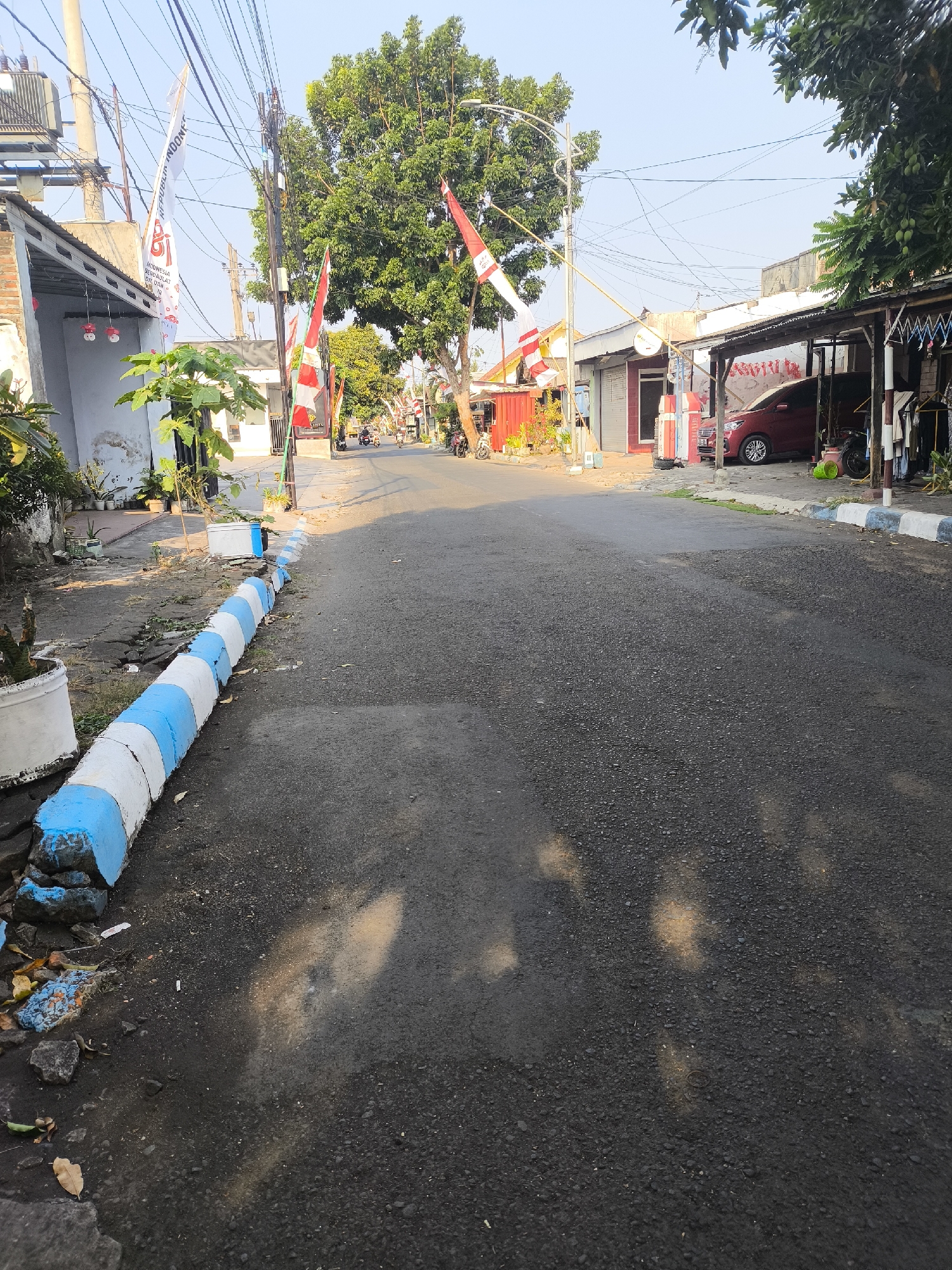 The image depicts a street scene in the city of Citrodiwangsan, Indonesia. The street is paved with asphalt and runs through a residential or commercial area. On the left side of the street, there is a row of houses and small shops, with some potted plants and a white and blue painted curb. Indonesian flags, red and white, are visible on poles along the street, suggesting a national celebration or event. A large tree with green foliage stands in the middle ground, casting shadows on the road. On the right side of the street, more buildings and shops are visible, including a red car parked under a covered area. Several motorcycles are also present, and a few individuals are seen riding them in the background, indicating ongoing activity in the area. The sky is clear and bright, suggesting it is daytime. The overall impression is of a typical street in an Indonesian town, possibly decorated for a special occasion.