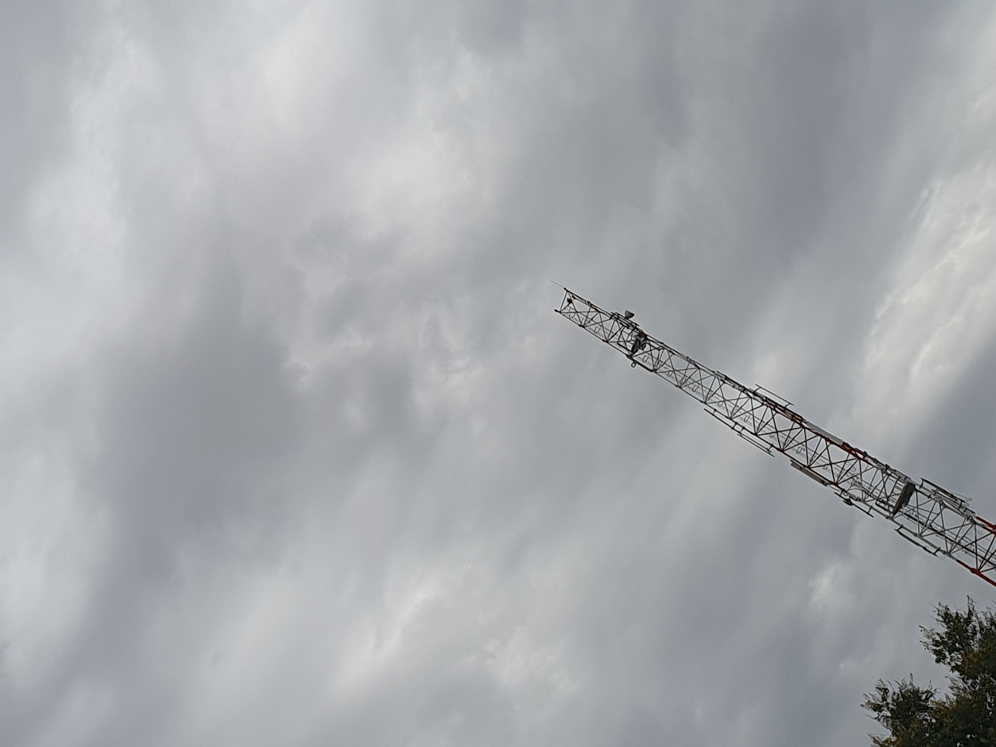 The image depicts a tall, latticed metal tower extending diagonally upwards into a cloudy, overcast sky. The tower is red and white, with a lightning rod at its apex. At the bottom right of the image, the tops of green trees are visible. The image was taken in the city of Kuru Dungus, Nigeria.