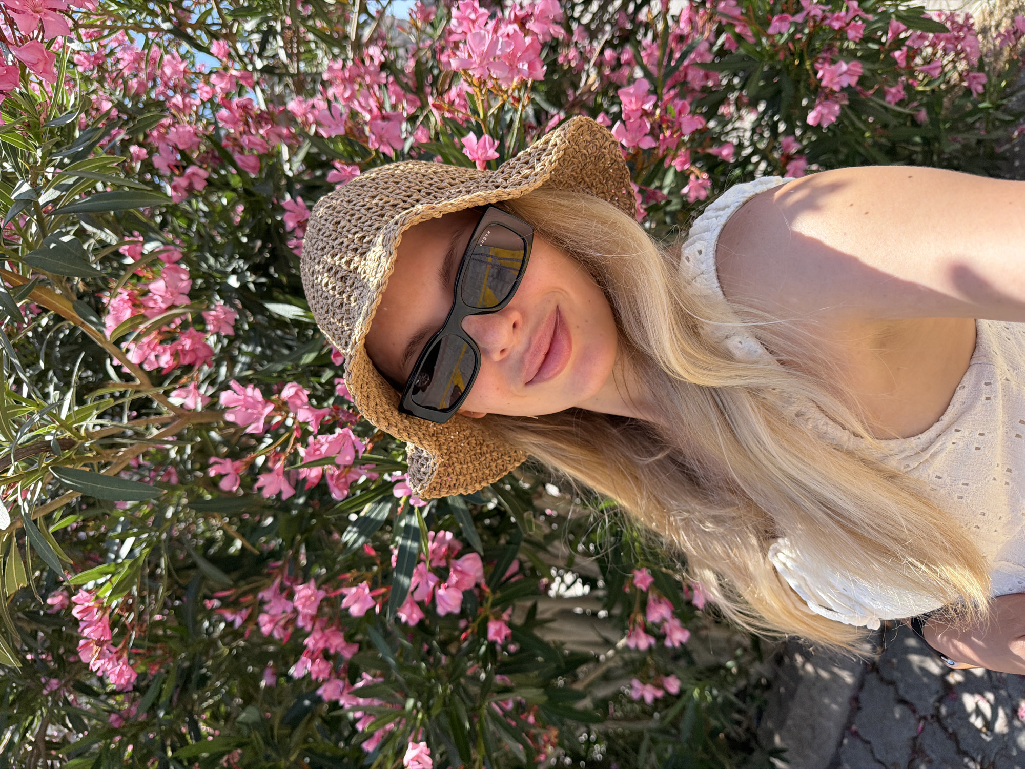 A blonde woman wearing a straw hat and sunglasses poses for a selfie in front of a flowering bush. The woman is smiling and looking at the camera. She is wearing a white top. The flowering bush is pink and green. The background is blurred, but it appears to be an outdoor setting. The woman's hair is long and blonde. The hat is woven and has a wide brim. The sunglasses are black and rectangular. The bush has many small pink flowers. The lighting is bright, suggesting it is daytime. The location is not explicitly stated in the image or available context.
