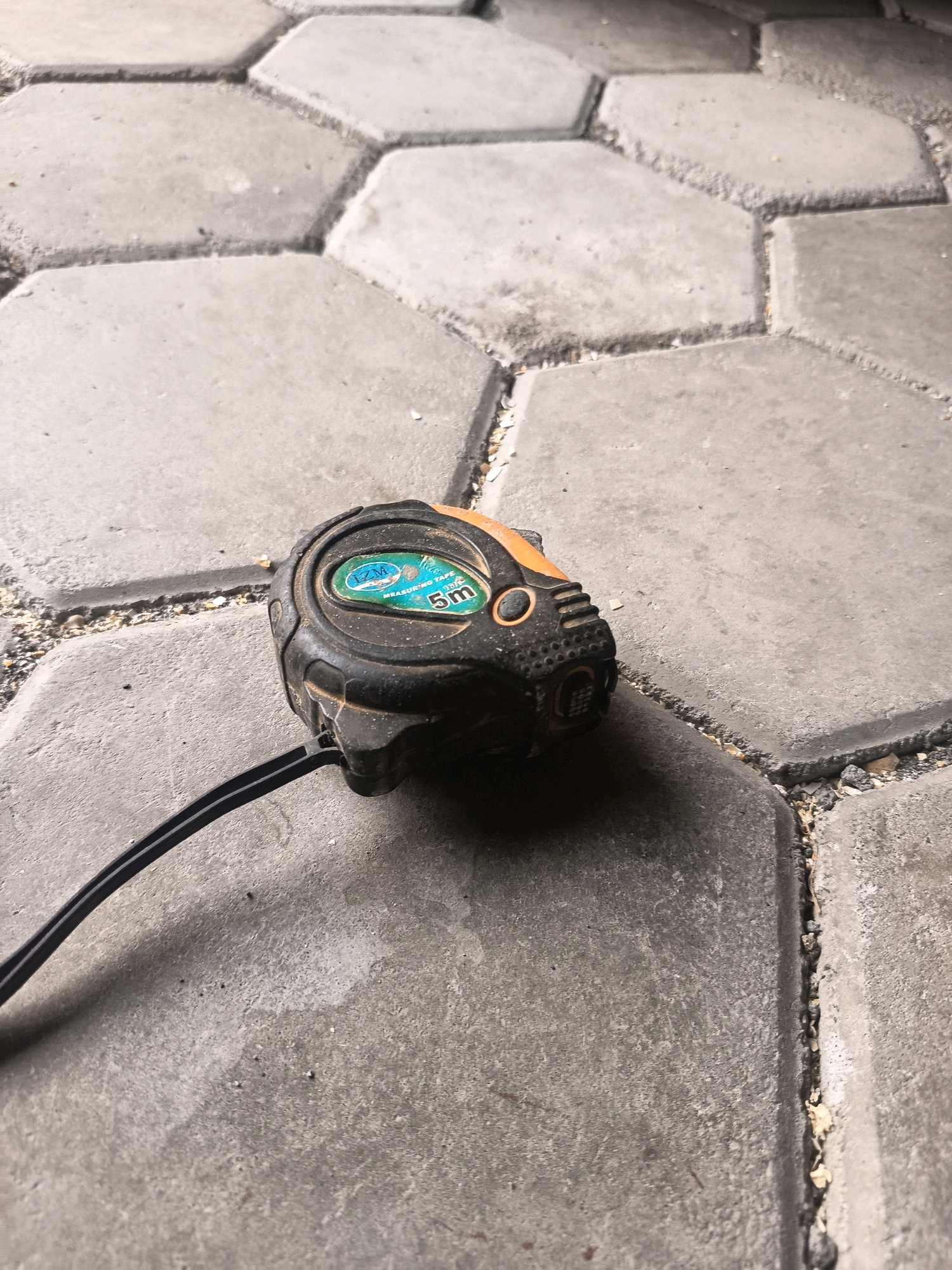 A tape measure rests on hexagonal pavers in Sidorejo, Indonesia. The tape measure is black with orange accents and has a blue and green label indicating "IZM MEASURING TAPE 5m". A black cord extends from the tape measure and lies on the pavers. The pavers are laid out in a honeycomb pattern and are a light gray color. There is some debris and sand between the pavers. The lighting suggests it is daytime.