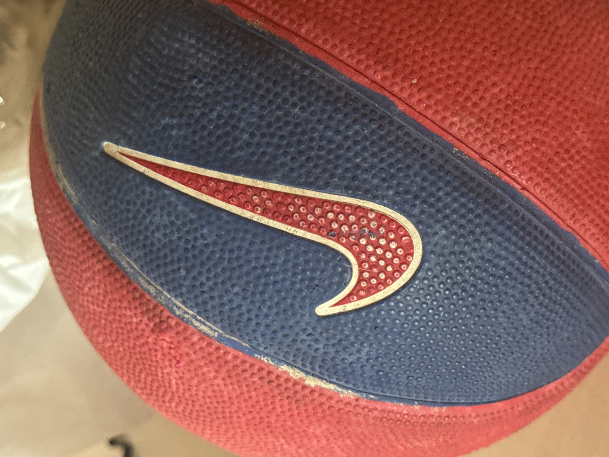 A close-up, high-angle shot of a red and blue Nike basketball. The basketball is textured with small bumps, and the Nike swoosh logo is prominently displayed in red with a white outline. The background is blurred and out of focus, but appears to be a neutral, indoor setting. There are no people or other distinguishing environmental features visible.
