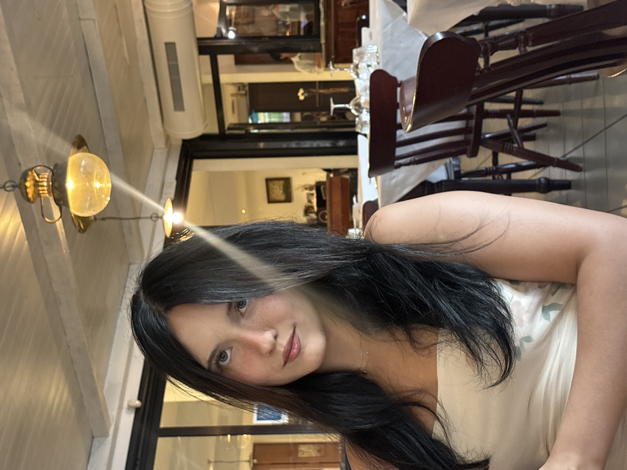 The image shows a young woman with long, dark hair and light skin with freckles, wearing a cream-colored strapless top. She is looking upwards and to the right. The background depicts a restaurant setting with tables covered in white tablecloths, set with glassware. Dark wooden chairs are visible around the tables. The ceiling has exposed wooden beams and decorative hanging lights. The lighting is warm and atmospheric. In the background, a person in a light-colored shirt is visible through a doorway. This scene is set in a restaurant, possibly in San Benedetto del Tronto, Italy.