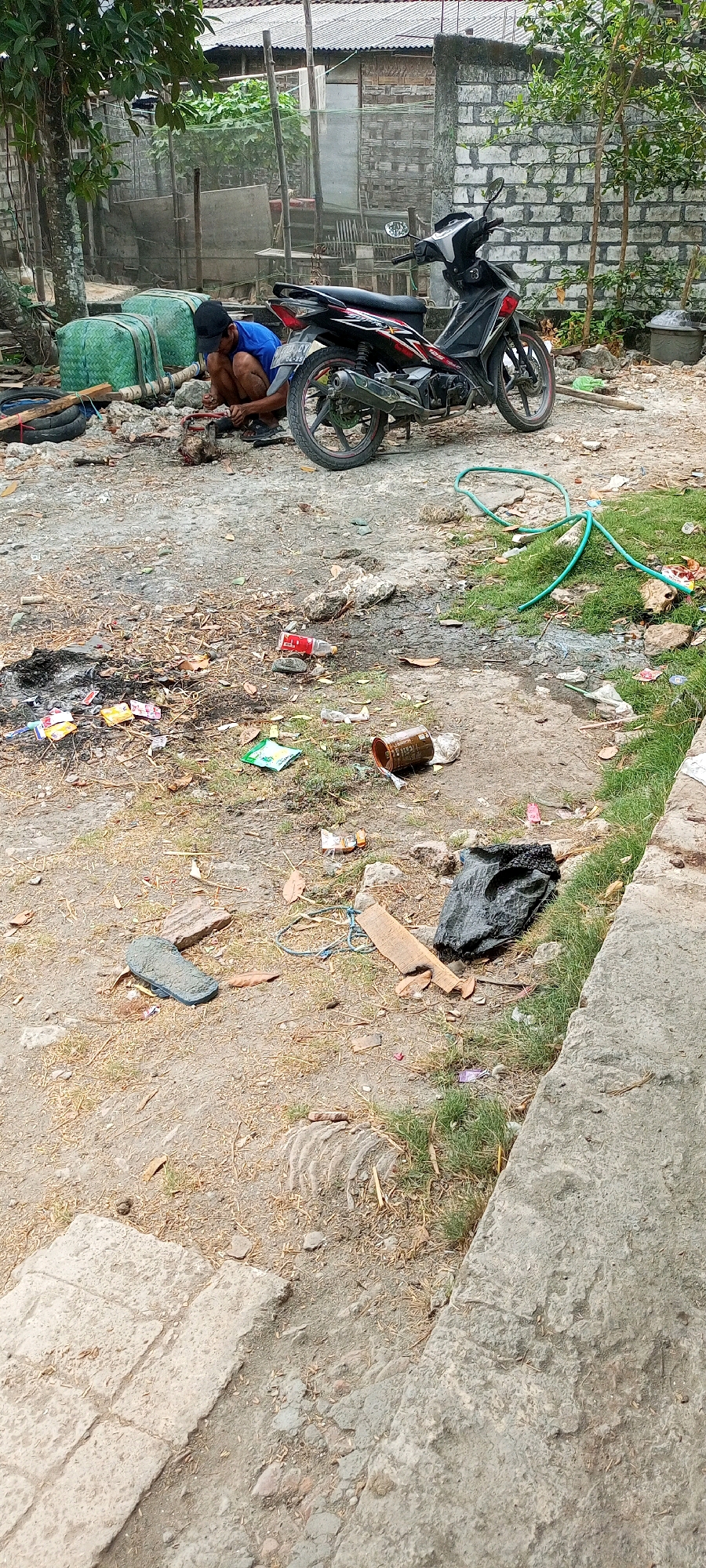The image depicts an outdoor scene in the city of Sambangan, Indonesia, characterized by a cluttered and somewhat disheveled environment. In the middle ground, a man wearing a blue shirt and a dark cap is crouched down, appearing to be engaged in some form of manual work. He is positioned near a parked motorcycle, which is predominantly black with red accents. The ground is a mixture of dirt, gravel, and scattered debris, including plastic bags, wrappers, a discarded can, and what appears to be burnt material, suggesting a fire may have occurred. A green garden hose is visible on the ground in the middle right of the frame, and to the left, several large green-wrapped bundles are present. Further in the background, there are structures including a building with a tiled roof and walls made of what looks like brick and wood. The foreground features a concrete path or ledge, with more dirt and sparse vegetation. A single sandal is also visible on the ground in the lower left quadrant.