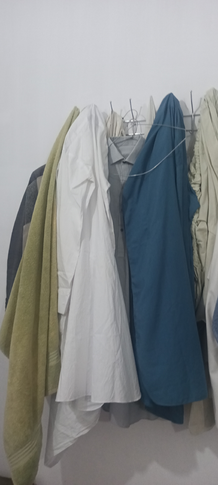 The image displays a collection of clothing items hanging on a rack against a white wall. The clothing includes a light green towel, a white shirt, a gray collared shirt, and a blue garment. Some of the items are draped over hangers, while others appear to be draped or folded. The lighting suggests an indoor environment, possibly a closet or a laundry area. The context provided places this scene in Peshawar City Tehsil, Pakistan.