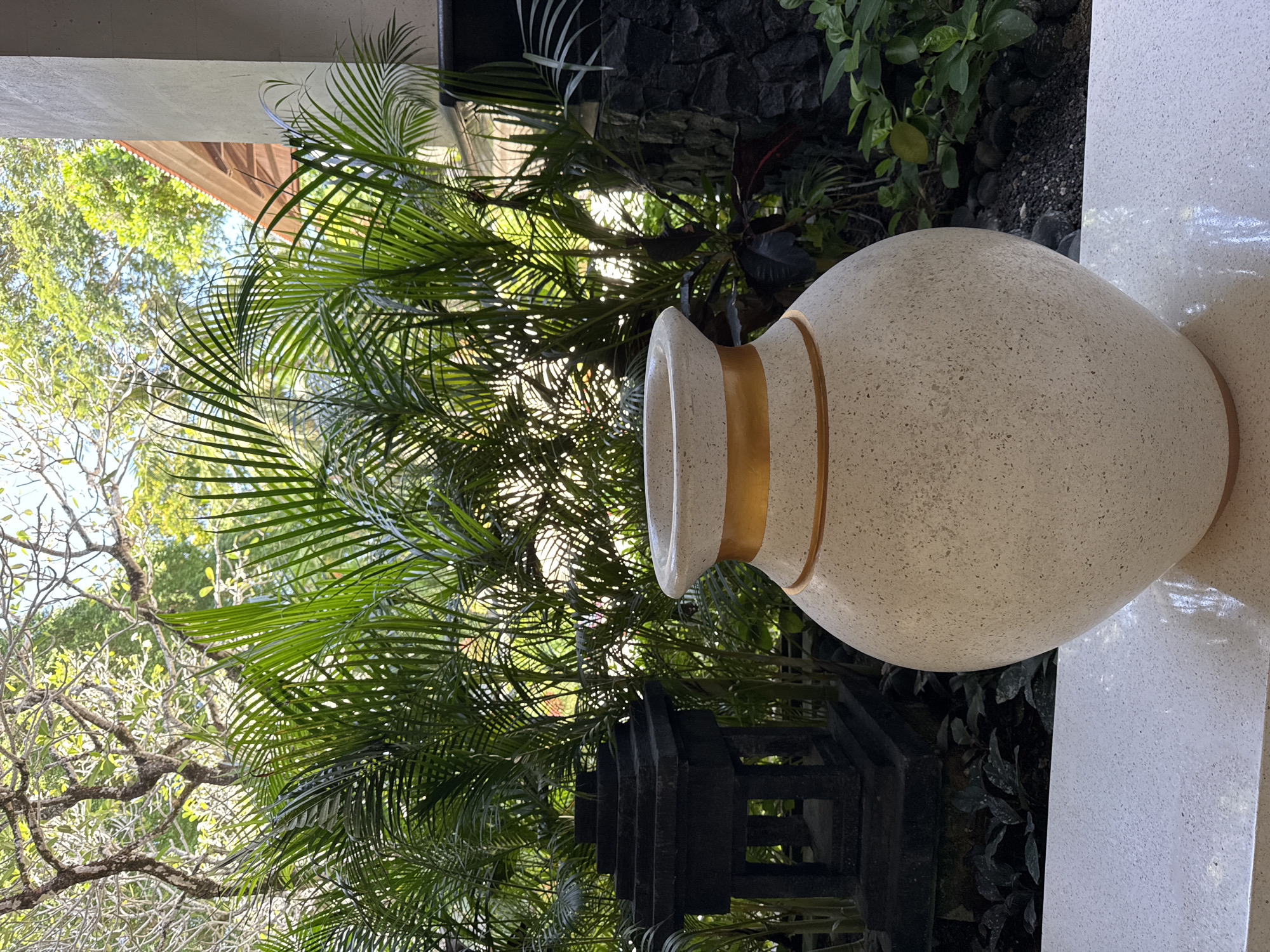 A close-up outdoor shot shows a large, light-colored stone or ceramic vase with a gold band around the neck, positioned near lush tropical foliage. The greenery includes palm fronds and other plants, providing a natural backdrop. A portion of a white column is visible on the right side of the frame, suggesting a building's exterior. The image appears to be taken during the day, with natural light illuminating the scene. The specific location is Kuta Selatan, Indonesia. A dark-colored architectural element is also present, possibly part of a fence or decorative structure.