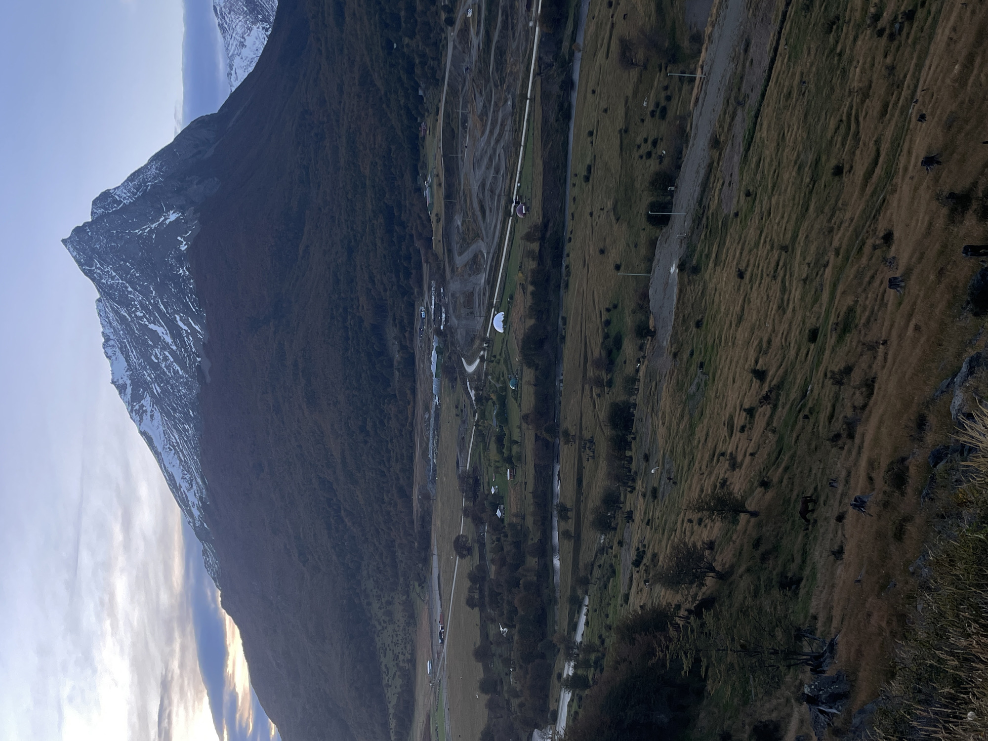 The image depicts a mountainous landscape in what could be Ushuaia, Argentina, though definitive Ushuaia landmarks are absent. A large mountain with a snow-capped peak dominates the left side of the frame. The mountain slopes down to a valley containing sparse vegetation, small buildings, roads, and what appears to be a river or stream. The foreground shows a hillside with grassy vegetation. The sky is visible above the mountain, with clouds. No people are distinctly visible. The orientation of the image is rotated.