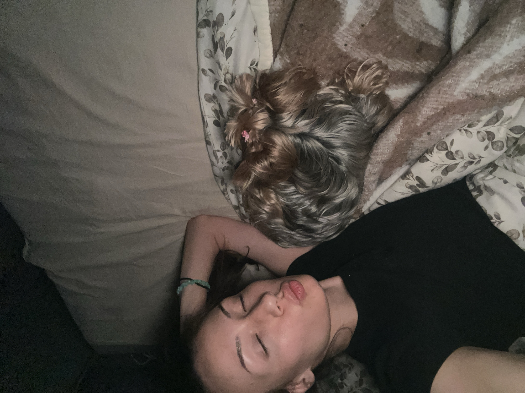 A woman and a small dog are lying in a bed. The woman is lying on her back with her eyes closed and her arm bent and resting beside her head. She is wearing a black top and a bracelet on her wrist. The dog is lying on its side near the woman's head. The bed is covered with sheets and a blanket. The sheets have a simple leaf pattern, and the blanket is brown with a plaid pattern. The scene is indoors. There is no indication that the photo was taken in Burgas, Bulgaria.