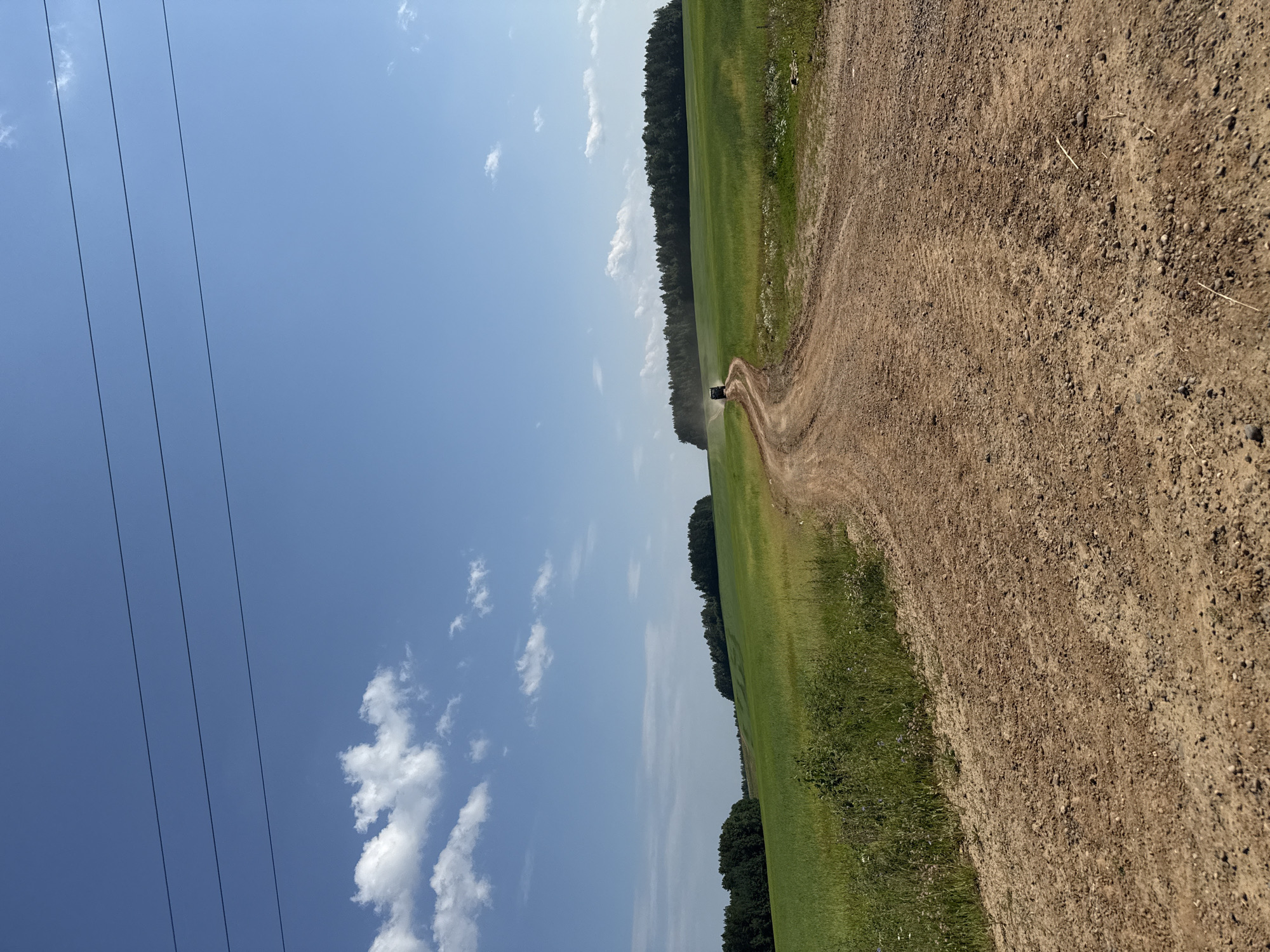 The image depicts a dirt road winding through a grassy field on a sunny day. The sky is a clear blue with scattered white clouds. Power lines are visible on the left side of the image. A dark, agricultural vehicle is driving on the dirt road in the distance. The field is bordered by a dense line of trees. The scene appears to be rural, possibly in Бабка, Russia, given the context. The time of day appears to be daytime, and the weather is sunny and fair. There is no visible text in the image.