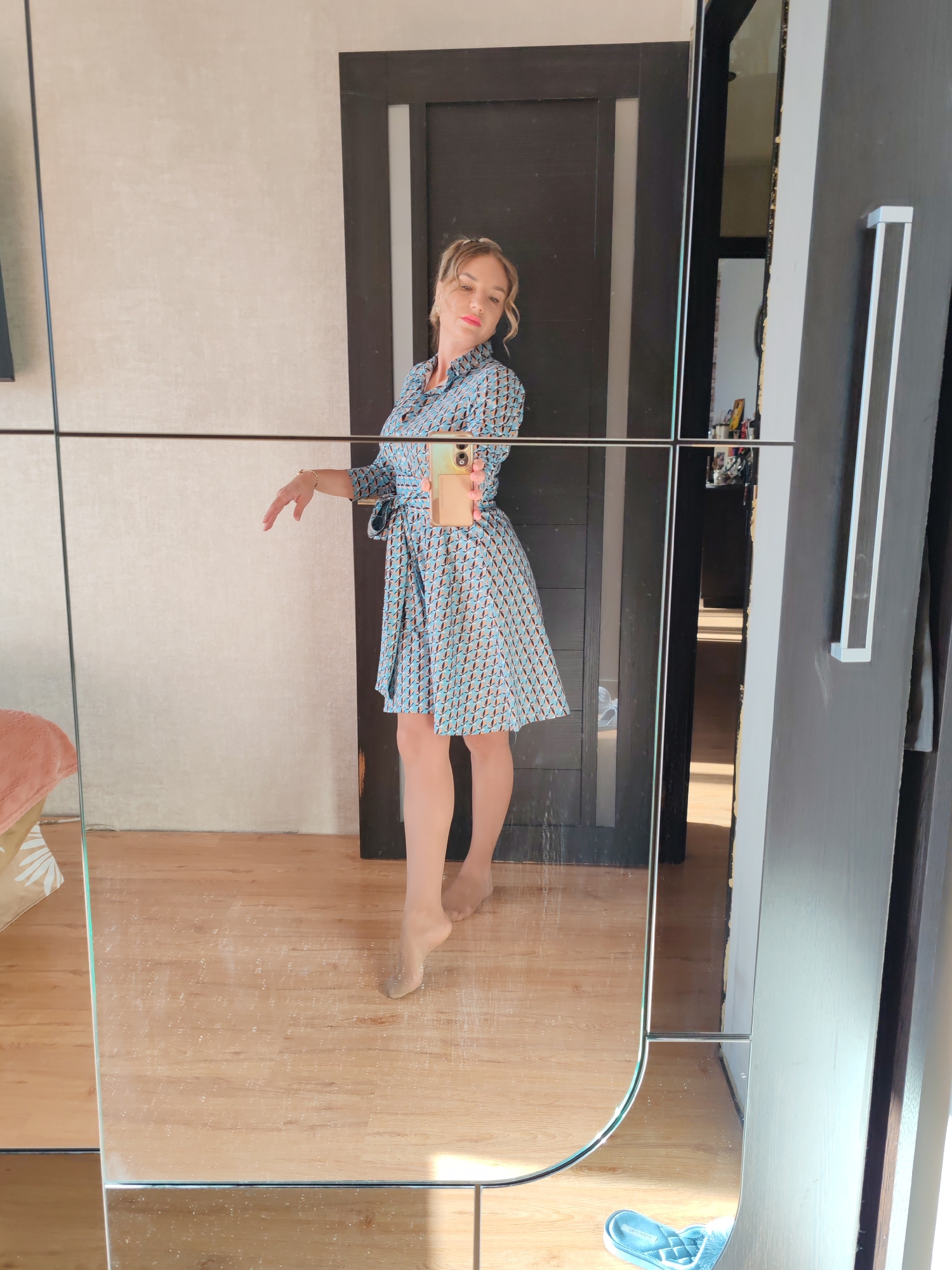 A woman is standing in a room, seemingly posing for a photograph in front of a mirrored wardrobe. The wardrobe has sliding doors with dark frames and silver handles. The woman is wearing a light blue dress with a geometric pattern and a tie belt. She's holding a smartphone in her right hand, presumably taking the photo. She appears to be wearing sheer stockings. The room has wooden flooring and plain walls. A partially visible bed with a patterned headboard is in the left corner. In the reflection of the mirror, a doorway and a pair of blue slippers on the floor are visible. The exact location in Petropavlovsk-Kamchatsky, Russia, is not discernible from the image.