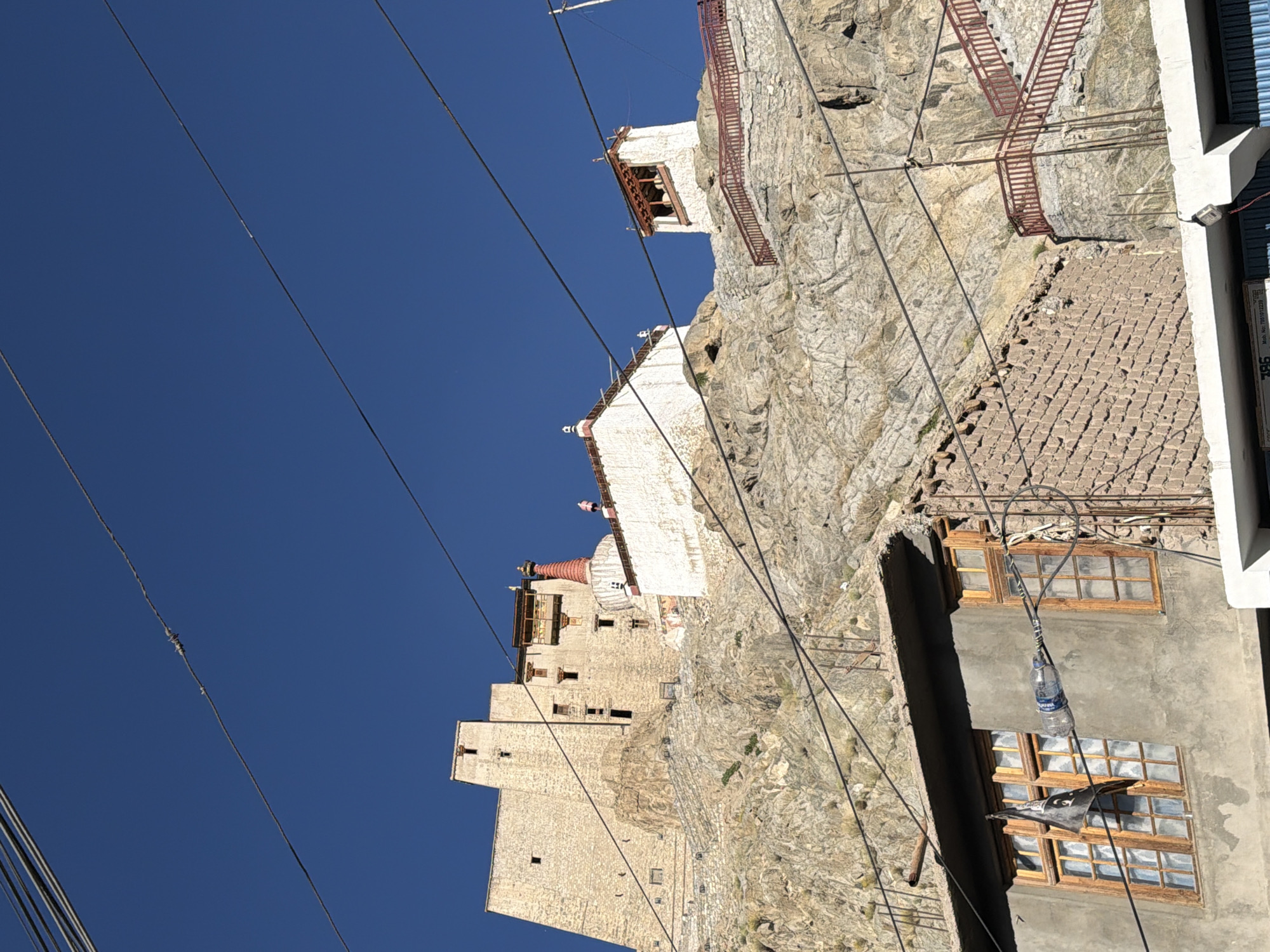 This is a low-angle, outdoor, daytime, vertical shot of the Leh Palace complex in Leh, India. The clear blue sky serves as the backdrop for a series of power lines that stretch across the frame. Dominating the mid-ground is the Leh Palace, a multi-storied, sandstone structure built into a rocky hillside. Various white and red-roofed structures, possibly monasteries or stupas, are interspersed with the palace's architecture. A red-railed staircase ascends the rocky slope towards one of the buildings. In the foreground, a section of a residential building with multiple windows and a grey concrete facade is visible, with more power lines and a plastic bottle hanging from one of them. The environment is dry and rocky, characteristic of the high-altitude desert climate of Ladakh. No people are clearly visible.
