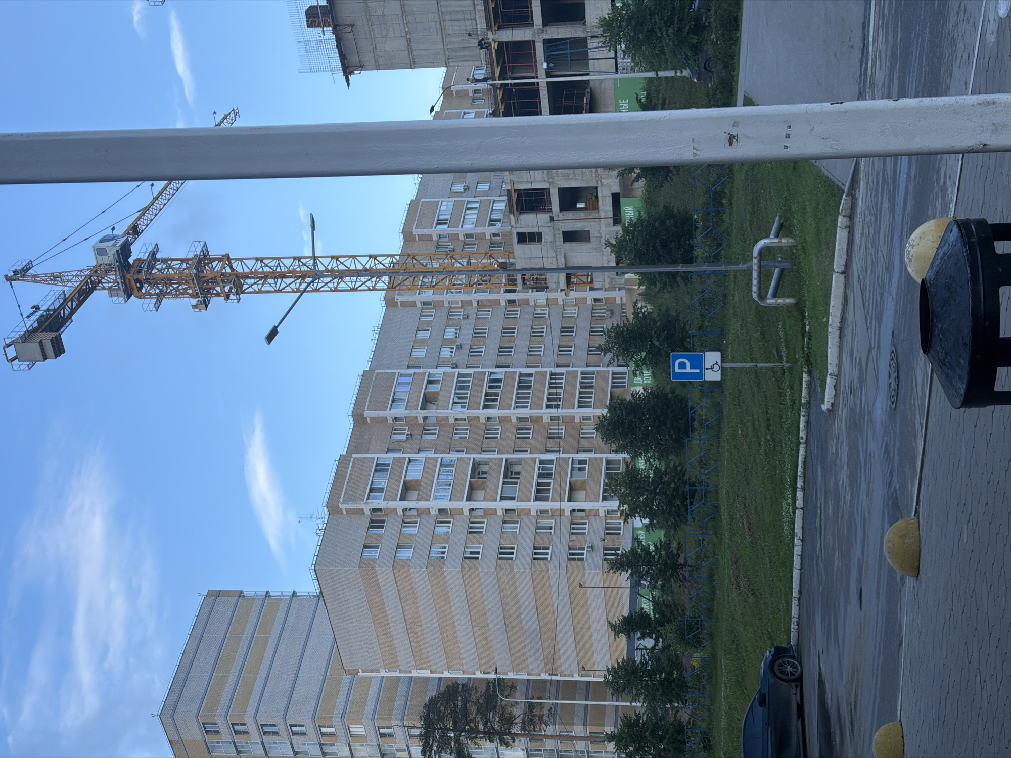 The image depicts an exterior view of an urban construction site in the city of Perm, Russia. A tall, yellow construction crane dominates the skyline, positioned behind a row of apartment buildings. The buildings themselves are a mix of completed structures and partially constructed ones, indicating ongoing development. One completed apartment building on the left is tall and modern, with horizontal bands of grey and beige brick. Next to it, a longer apartment building features a similar brick pattern and numerous windows. In the foreground, a paved area with a dark car parked on the left suggests a street or parking lot. Yellow bollards are visible, and a trash receptacle is present. A blue sign with a white 'P' and a wheelchair symbol indicates accessible parking. Green trees and shrubs line the area in front of the apartment buildings, and a blue fence runs along the edge of the property. The sky is a pale blue with scattered clouds, suggesting a daytime scene.