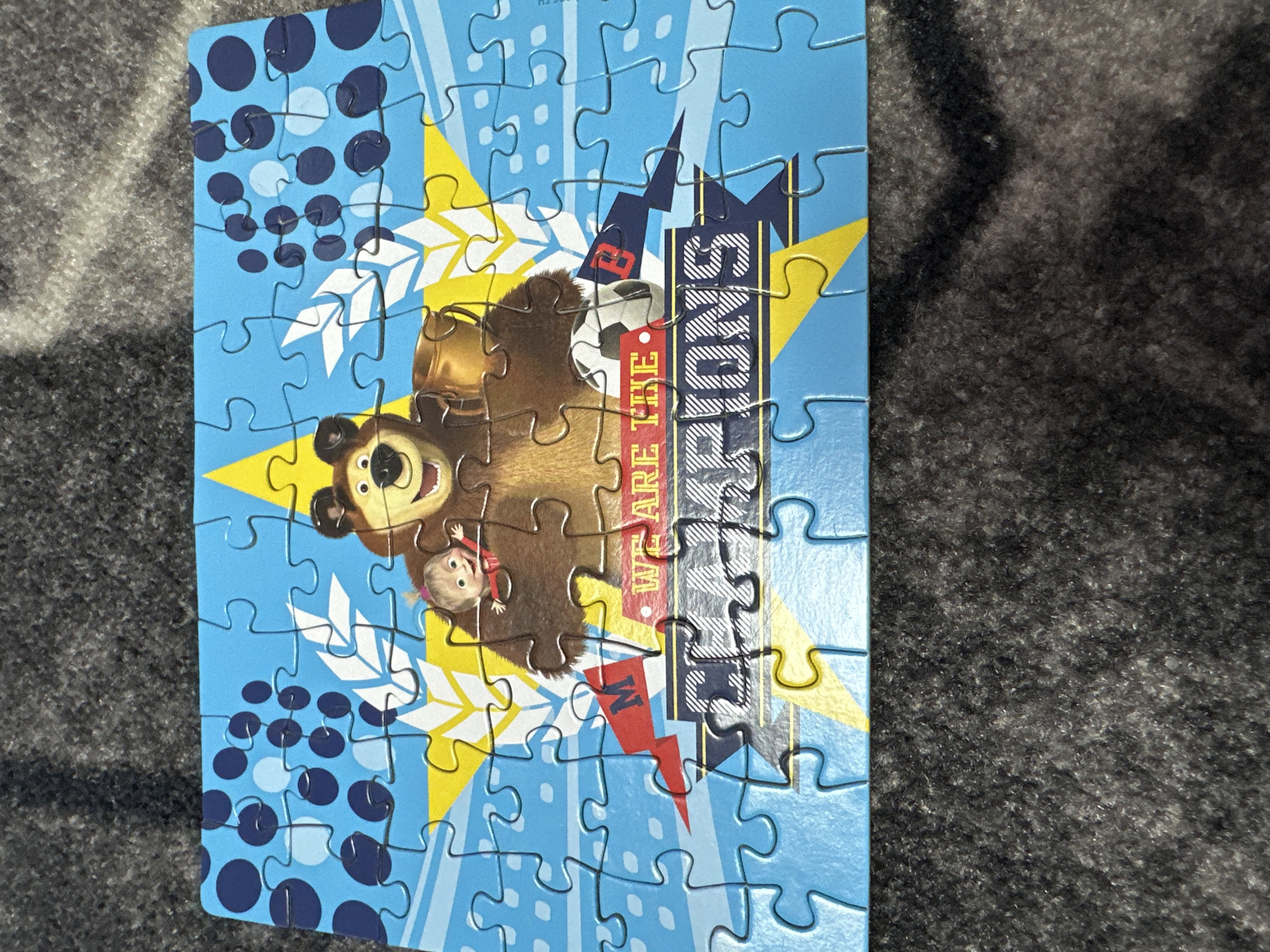 The image shows a completed jigsaw puzzle depicting Masha and the Bear. The background is light blue with blue circles and yellow lightning bolts. The text "WE ARE THE CHAMPIONS" is visible on a gray striped banner. A soccer ball with the number 8 is near the bear.
The location of Chaikovsky, Russia, is not depicted in the image.