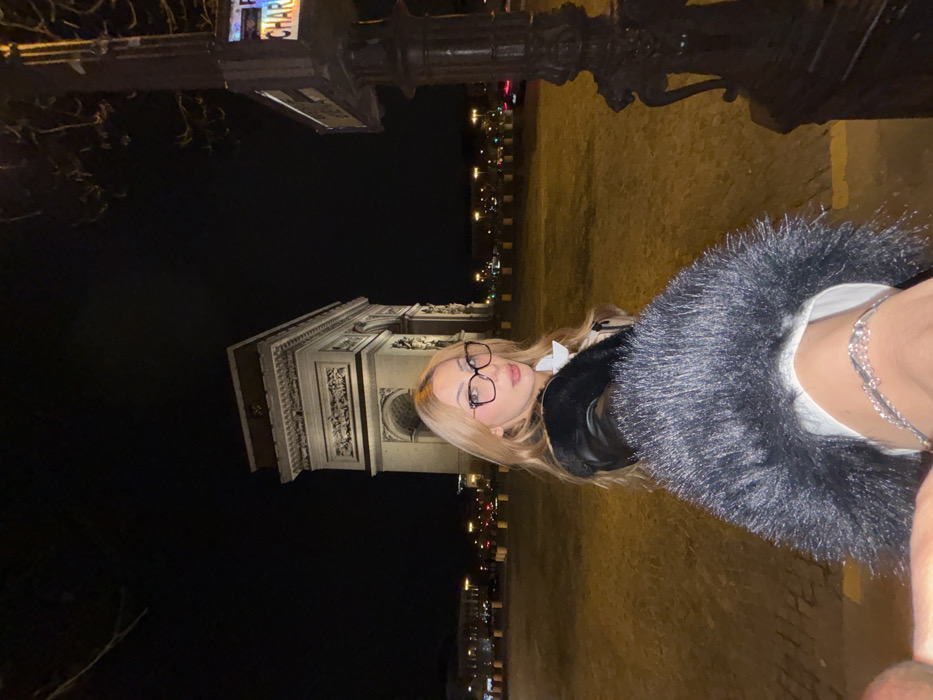 The image depicts a nighttime scene in Paris, France, with the Arc de Triomphe prominently displayed in the background. A female individual occupies the foreground, looking directly at the camera. She has long blonde hair and wears dark-framed eyeglasses. Her attire includes a black jacket with a fur or faux-fur trim, over a white garment. A silvery, multi-strand chain accessory is visible across her upper right thigh. Her right arm extends towards the camera, which is positioned at a low angle. The ground in the mid-ground is illuminated, appearing yellowish. To the far right, a dark lamppost with a street sign partially visible, displaying "CHARL," stands. City lights and traffic are discernible in the distant background, and bare tree branches are visible against the dark sky at the top.