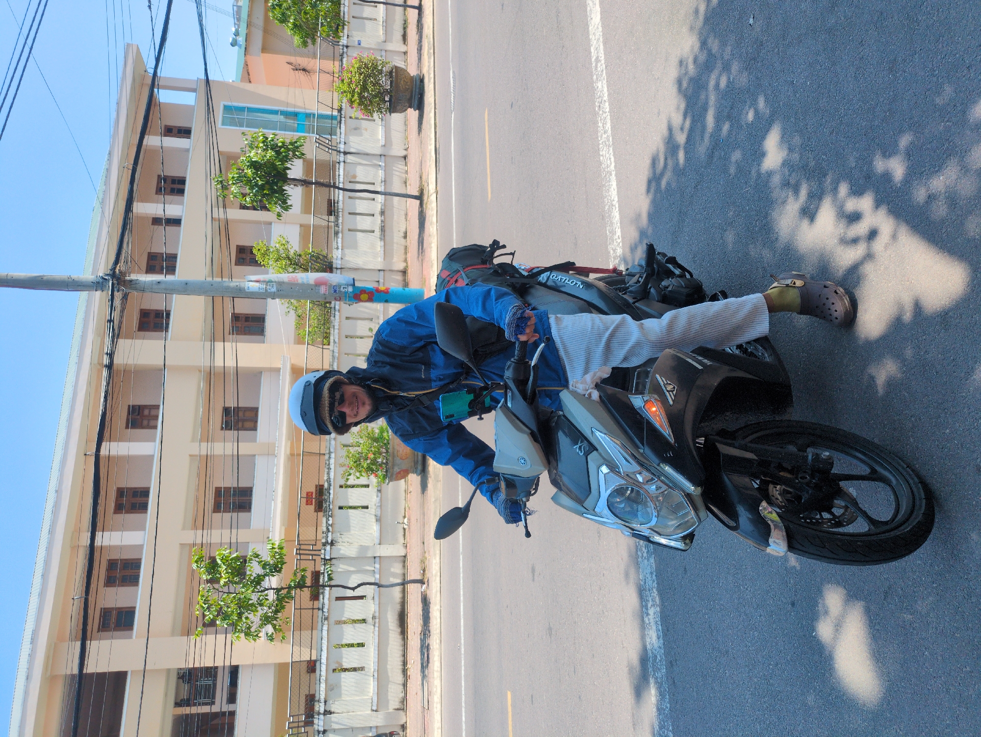 A person wearing a blue jacket, white helmet, and sunglasses is sitting on a motorcycle on a paved road in South Quy Nhon Ward, Vietnam. The person is wearing light-colored pants and brown shoes with holes. A building with multiple windows and a fence runs along the left side of the road. There are trees planted along the fence. The road has a white line marking lanes. A utility pole is visible near the building.