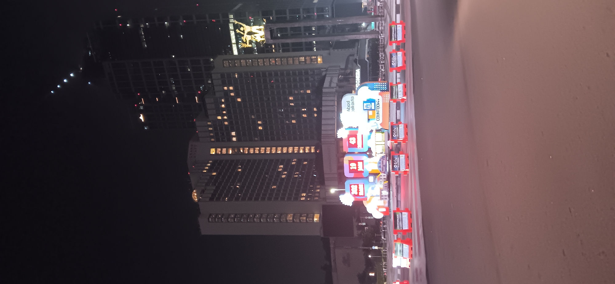 At night in Central Jakarta, Indonesia, the intersection is illuminated by the bright lights of several skyscrapers, including the Grand Hyatt. In the foreground, a colorful, illuminated display features large numbers "302", "10", and "43" on screens, along with the words "Abadi Jakarta" and "COUNTDOWN". Orange and white traffic barriers are lined up on the road, some displaying logos of "OTRIV" and "CRYPTOWAVE". The overall scene is a bustling urban environment at night, with a prominent festive or event-related display.