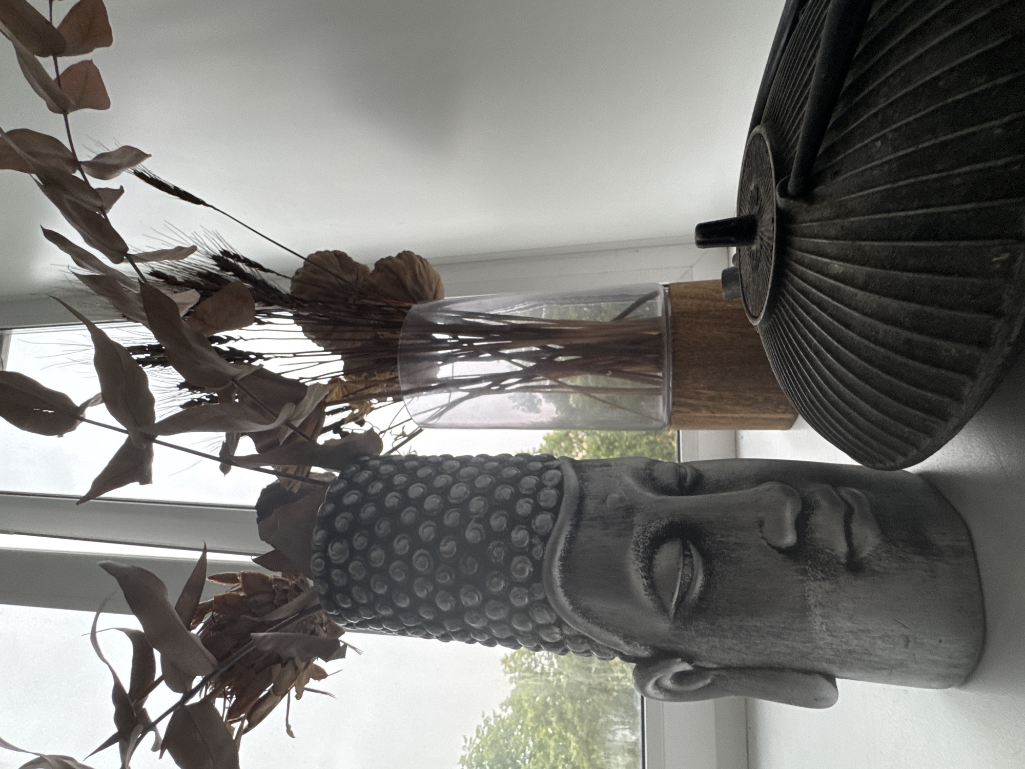 The image depicts a still life arrangement on a windowsill. In the foreground, a dark, ribbed metal object, possibly a teapot or decorative lid, is positioned to the right. To its left is a grey, Buddha head-shaped vase filled with dried floral arrangements, featuring brown leaves and stalks. Behind the Buddha vase, a clear glass vase with a wooden base holds similar dried plants. The objects are placed on a white surface, likely a windowsill, with a window providing a backdrop of greenery from trees outside and a bright, overcast sky. The overall scene is indoors, with a plain white wall on the right side. There are no people or animals visible in the image. The lighting is diffused, suggesting an overcast day. No specific location in Moscow, Russia can be determined from the visual information.