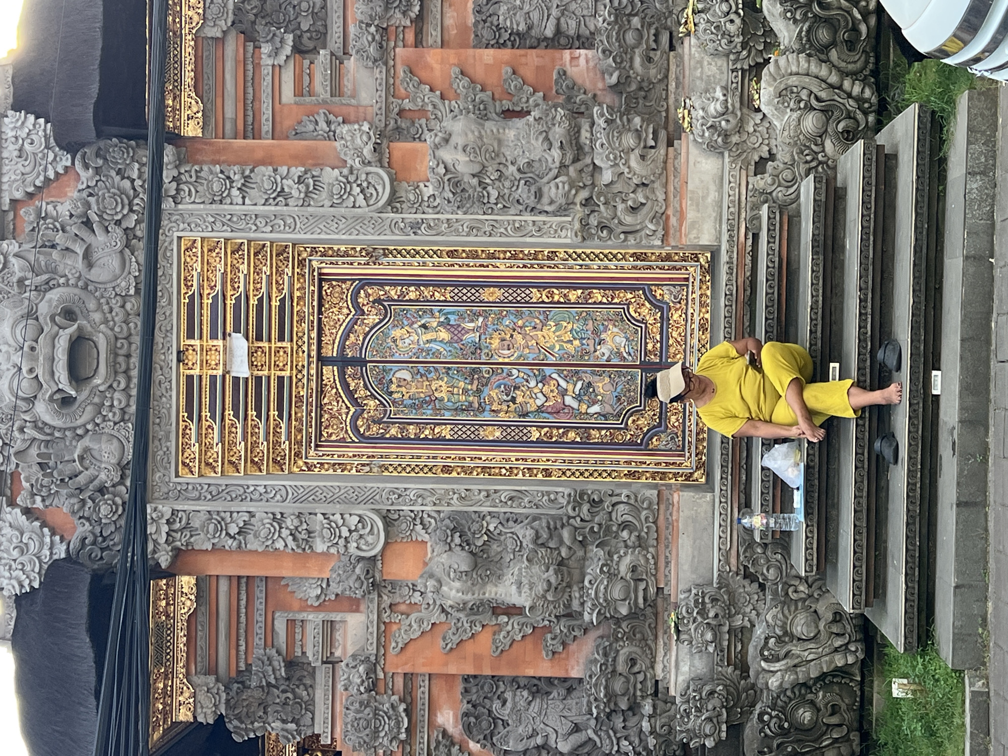 A medium-shot, rotated image depicts a person resting on the steps of a building decorated with intricate carvings, likely in the Ubud District, Indonesia. The person is sitting on the steps, facing right, and dressed in a yellow outfit with a white cap. Their shoes are placed next to them. A water bottle and a plastic bag are beside the person on the steps. The backdrop features ornate stone carvings of mythical figures and floral patterns. A decorated, rectangular structure with gold and dark blue trim and intricate artwork, possibly a door or a wall panel, is centrally placed in the background. The architecture and carvings suggest a cultural or religious site, potentially a temple.