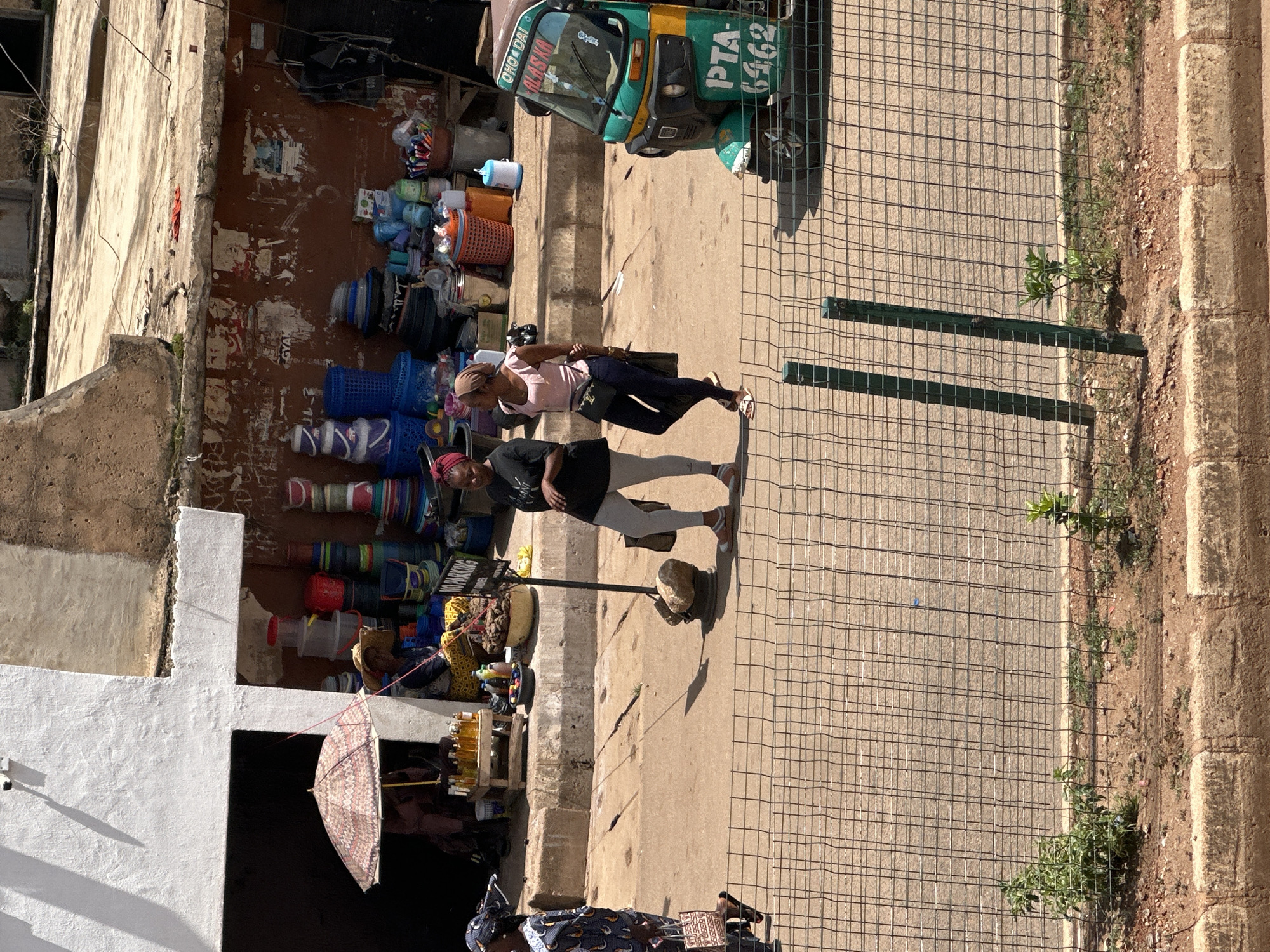 The image depicts a bustling market scene in Bukuru, Nigeria, viewed from a slightly elevated perspective. The time of day appears to be daytime, with strong sunlight casting distinct shadows. The weather seems clear and warm, typical of a sunny day.
In the foreground, two women are the primary subjects. One woman, wearing a pink top and dark leggings, is walking, carrying a dark bag over her shoulder. Another woman, in a black top, is walking ahead of her. The general atmosphere seems to be that of everyday commerce and movement.
The setting is an outdoor market adjacent to a building with weathered walls, suggesting an established commercial area. A variety of objects are visible, including stacked plastic containers and baskets of different colors and sizes, indicating merchandise for sale. A prominent object is a brightly colored auto-rickshaw, a common mode of transport in Nigeria, parked nearby. This auto-rickshaw has text on it, including "OHO DAI" and "PTA 6462".
Further into the scene, there are stalls or displays with more items, including bottles of liquids, possibly oils or beverages, and a patterned umbrella. The ground is paved, with a section of gravel and vegetation along the right edge, bordering a low wall made of concrete blocks. A wire fence runs along the pathway, separating the market area from the street or sidewalk.
A small sign is visible with the text "NO PARKING". The overall impression is one of a vibrant, active marketplace, typical of many urban centers in Nigeria. The image captures a moment of daily life and economic activity.