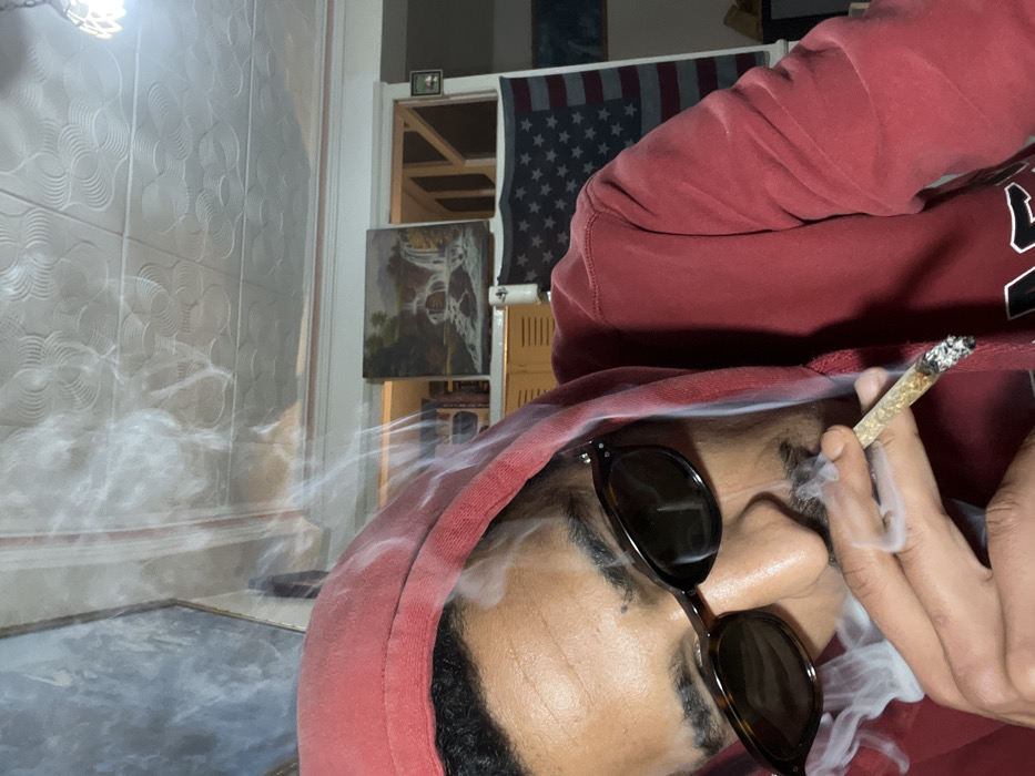 An individual is depicted indoors, in what is stated to be the city of Tanger, Morocco, in a close-up, eye-level shot. The person, appearing male and wearing dark tortoise-shell sunglasses and a red hooded sweatshirt, is holding a lit, hand-rolled cigarette (joint) between the thumb and index finger of their right hand. Smoke is actively rising from the joint and from the area around the individual's mouth and nose, indicating recent inhalation or exhalation. The background reveals an interior space with a white, patterned ceiling. Behind the individual, a painting depicting a waterfall scene is visible above a shelf or cabinet. To the right, an American flag is draped, and further right, wooden structural elements are discernible. Another large, possibly abstract, artwork is partially visible on the far left.