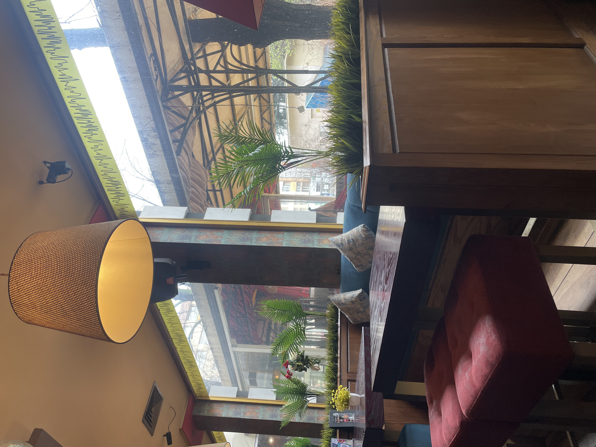 The image captures an indoor scene, seemingly in a cafe or restaurant in Varna, Bulgaria, suggested by the architecture and overall style. The perspective is slightly tilted, showcasing the interior and a glimpse of the outside.
The main elements visible are:
- **Interior**: A large lampshade hangs from the ceiling, providing warm light. There are seating arrangements with a red velvet bench and a dark-colored table, positioned next to a blue-upholstered bench. Pillows are scattered on the blue bench. There is also a wooden partition or wall with paneling.
- **Exterior view**: A window allows a view of a covered outdoor area with a metal structure and some vegetation. Buildings are visible in the background, indicating an urban environment.
- **Decorative elements**: Greenery, including potted plants and decorative grass, adds a touch of nature to the interior. There is a small vase with yellow flowers on the table.
- **Architecture**: The building's architecture is visible through the window, showing a mix of styles and potentially an older building.
- **Other**: A yellow strip with patterns is placed along the top of the window.
The scene conveys a sense of coziness and relaxation, typical of a cafe or restaurant setting.