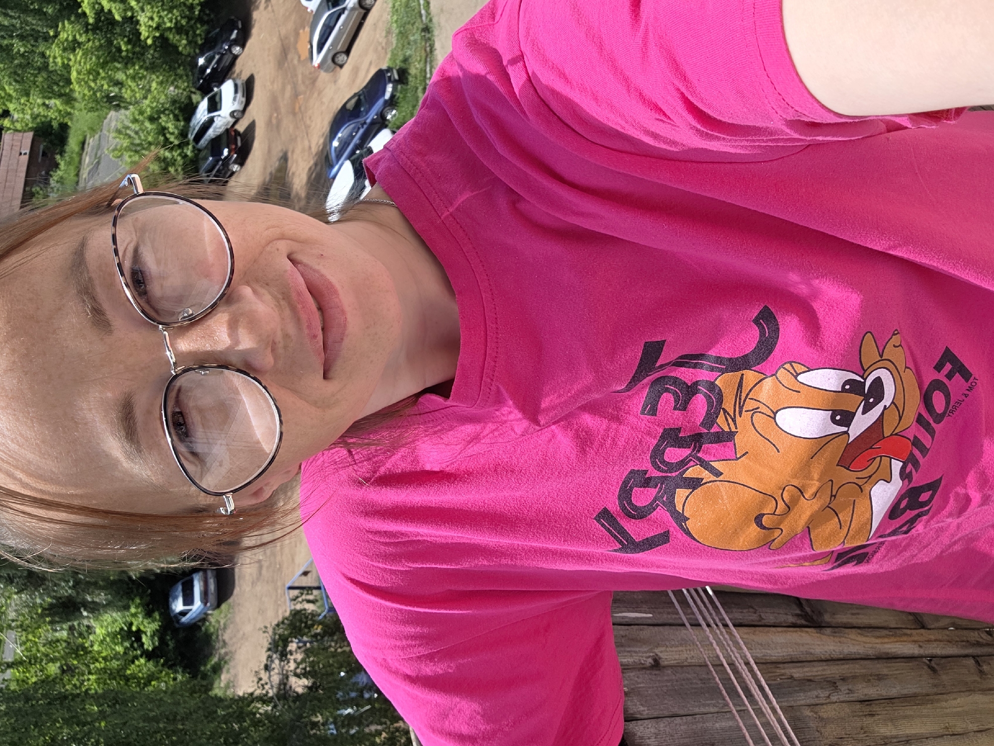 An image taken from an elevated position in Chaikovsky, Russia, features a woman in the foreground taking a selfie on a bright, sunny day. The woman has light reddish-brown hair, visible freckles across her nose and cheeks, and wears round, silver-rimmed eyeglasses. Her lips are slightly parted in a subtle smile. She is dressed in a vibrant pink t-shirt with a graphic print of the cartoon character Jerry holding an item, with the character's name "JERRY" printed above, appearing reversed in the selfie. The background reveals an outdoor environment. To the left, dense green foliage and trees are visible, with a light-colored sedan parked on a dirt surface. To the right, an unpaved parking area contains several vehicles, including multiple dark-colored sedans, a white minivan, a silver sedan, and a blue sedan. Beyond the parking area, additional green trees and a building with a reddish-brown roof are discernible in the distance. In the lower left foreground, a wooden barrier with several parallel wires or ropes is present.