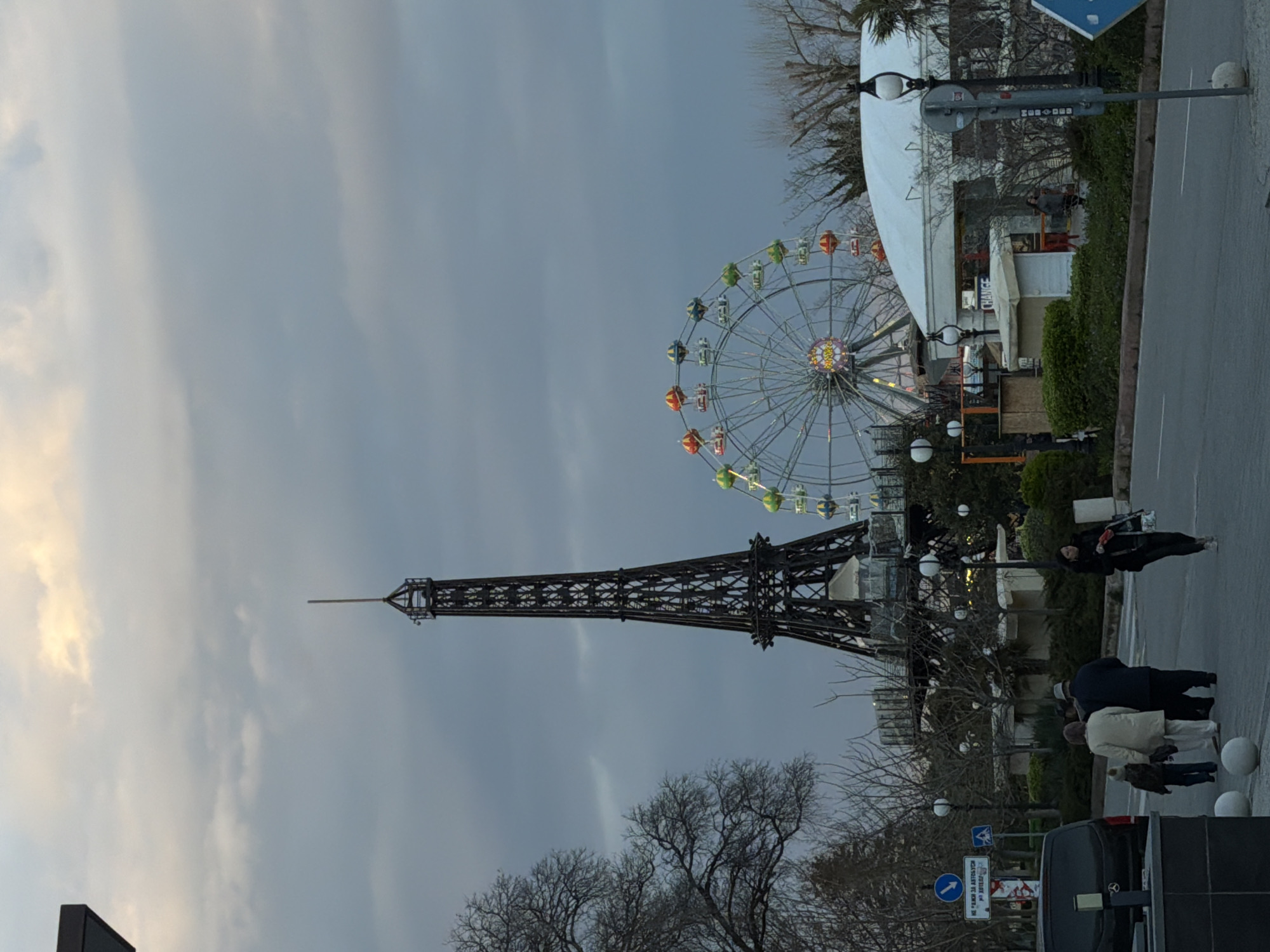 The image depicts a miniature Eiffel Tower and a Ferris wheel in Varna, Bulgaria. The sky is overcast. Several pedestrians are walking on the paved area adjacent to a street. Cars are parked nearby. Street signs are visible. There are leafless trees in the foreground. In the background is a white tent structure. Street lights line the pavement.