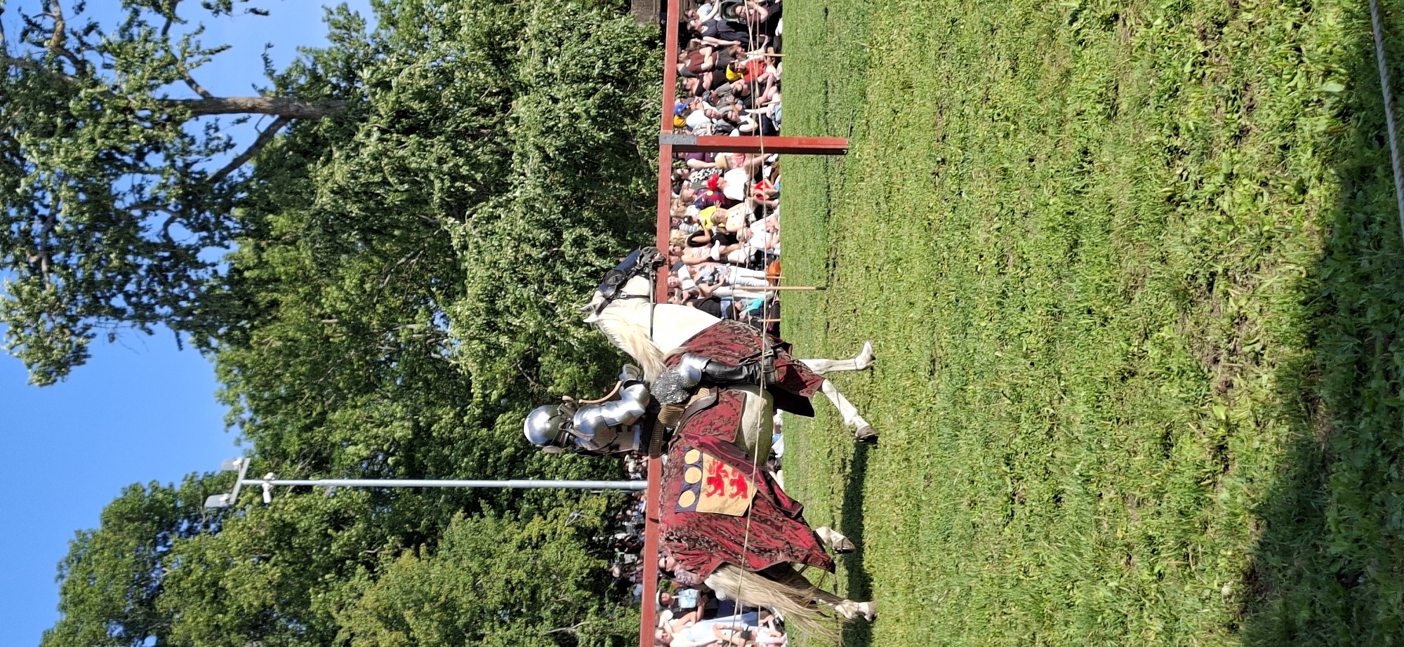 A person in full metal armor is riding a white horse across a grassy field. The horse is draped in a red and gold tapestry. A crowd of people are seated in the background, behind a red barrier. Trees and a bright blue sky are visible in the distance. This scene is likely from a jousting tournament or medieval reenactment event. The image does not provide specific information about the location within Turku, Finland.