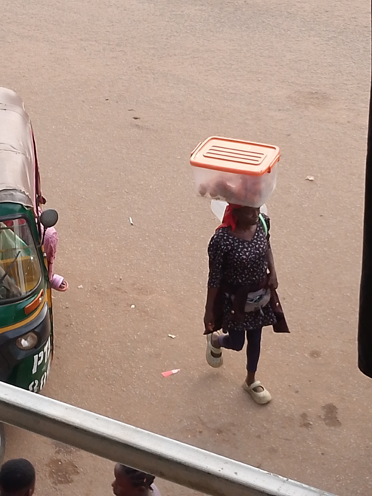 The image captures a street scene in Bukuru, Nigeria, from an elevated perspective, looking down onto a light brown or tan road. The main subject is a woman walking across the frame from right to left. She carries a clear plastic container with an orange lid balanced on her head, which appears to hold various items, possibly food. She wears a dark blue or black dress with a light-colored pattern, dark leggings, and light beige or off-white flat shoes. A red head covering is visible beneath the container, and a light-colored fanny pack or waist bag is slung around her waist.
To the left, a green and yellow tricycle, commonly known as a Keke Napep, is partially visible, either parked or moving slowly. A person's arm and a pink-striped sandal can be seen inside the vehicle. The letters "PTA" and some other unreadable characters are visible on the front of the tricycle. In the immediate foreground, a metal railing runs horizontally, suggesting the photo was taken from a balcony, window, or overpass. Below this railing, the tops of two heads, possibly children, are partially visible. The scene depicts typical daytime street activity, with soft natural light indicating it could be morning or late afternoon. The overall atmosphere is that of an ordinary day in an urban or semi-urban setting. The road surface shows minor debris or dirt in places.