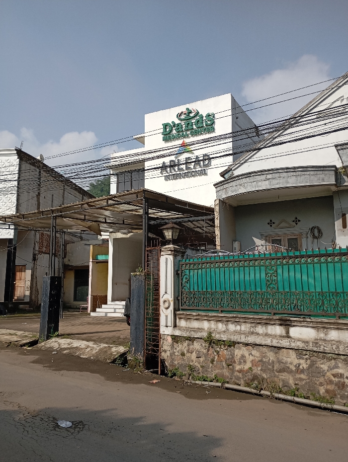 The image depicts a daytime street scene in the city of Situwangi, Indonesia. The central focus is a multi-story, white commercial building identified by prominent green signage that reads "Dian's Medical Center" with a cross emblem, and "ARLEAD INTERNATIONAL" in black lettering beneath. This building features multiple windows on its visible facade.
In the right foreground, a residential structure with a teal-colored, ornate metal fence atop a stone and concrete wall runs parallel to the street. An ornate white pillar, part of a gate structure, stands at the edge of the property line. Small weeds are visible growing along the base of this wall.
On the left side of the scene, several other buildings are present. One features a textured white facade, while another appears to have an unfinished or darker exterior. A metal framework structure with a corrugated roof or awning extends from one of these buildings towards the street. Power lines are visible crisscrossing overhead, connecting to poles not fully in frame and running across the sky and buildings.
The foreground consists of an asphalt road showing signs of wear, including cracks and some light debris. A darker, possibly unpainted or heavily weathered gate post is situated on the left side of the road, near the metal awning structure. The sky is largely clear blue with scattered white clouds, indicating fair weather and bright sunlight, which casts shadows from the fence and buildings. No people are present in the frame.