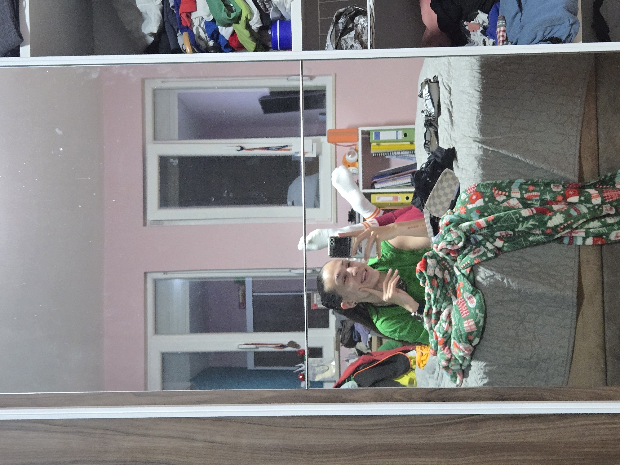 A mirrored wardrobe reflects a room in Pleven, Bulgaria. The reflection shows a young woman lying on a bed taking a selfie. She is wearing a green shirt, pink leggings, and white socks with orange stripes, and is partially covered by a green and red Christmas-themed blanket. The room has pink walls and a window. A bookcase with files and a white wardrobe with clothes are visible. The bed has a grey patterned cover.