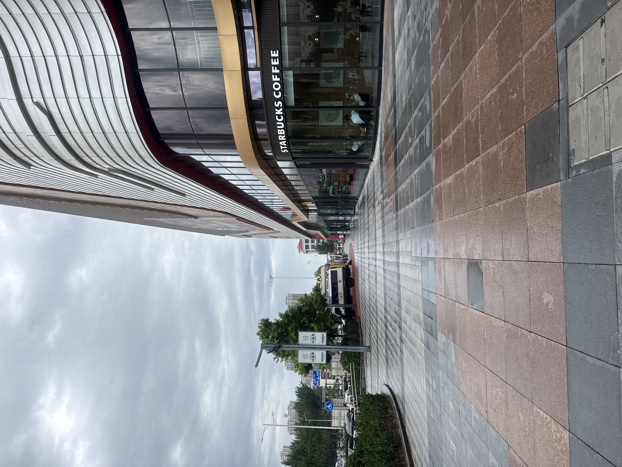 The image shows a wide, paved pedestrian walkway in Qinhuangdao, China, with a modern building on the right and a street with traffic on the left. The walkway is made of large square tiles in grey and reddish-brown, some of which appear wet, suggesting recent rain. On the right, the building features a curved facade with a metallic, undulating design. At street level, a Starbucks Coffee store is visible with large windows and patrons inside. Further down the street, various vehicles, including cars, a bus, and what appears to be a white van with "公安" (Public Security) written on it, are present. Trees line the left side of the street, with apartment buildings visible in the distance. The sky is overcast and grey.