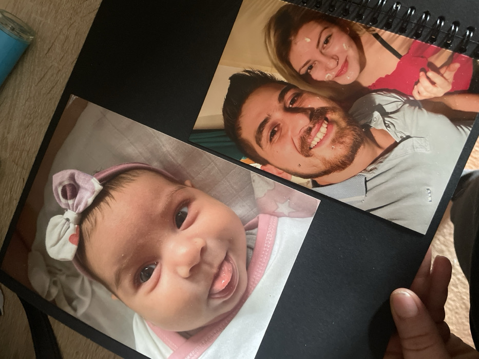 The image displays an open, coil-bound black photo album or scrapbook, viewed from an oblique angle. The album contains two glossy photographs. The upper photograph features a close-up of an infant with dark hair, wearing a pink headband adorned with a multi-patterned bow. The infant's eyes are open, and its tongue is extended slightly from its mouth. The lower photograph is a selfie-style portrait of two adults: a man and a woman. The man has a beard, dark hair, a lower lip piercing, and is smiling, wearing a light-colored polo shirt. The woman has blonde hair, a nose piercing, a white substance on her left cheek and chin, and is smiling while wearing a red top. Her left hand is visible, seemingly interacting with the man's arm. A human hand, with a visible thumb and fingers, is holding the lower-left edge of the album. The background behind the album appears to be a light-colored wooden surface, with a partial blue cylindrical object visible in the upper right corner. The specific location within Senden, Germany, cannot be determined from the visual content of this image.