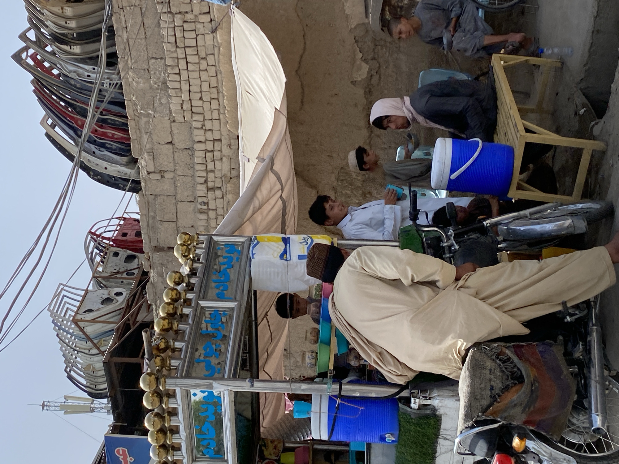 This is a photograph taken in the city of Kandahar, Afghanistan, capturing a scene that appears to be a street-side food or drink vendor, likely located in a market or busy area.
In the foreground, a man wearing a traditional beige tunic and a patterned skullcap is partially visible, leaning over the back of a motorcycle. The motorcycle has a colorful woven bag attached to its rear. Behind the man, and to his left, is a mobile cart which appears to be a vendor stall. It is decorated with golden spheres on top and has blue lettering on a sign that is likely in Dari or Pashto. Beneath the sign, there are blue and white containers, one of which is a large blue cooler.
Several young boys are present around the stall and in the midground. One boy, wearing a white shirt, stands near the stall, holding a blue cup. Another boy, wearing a grey tunic and a white skullcap, is partially visible behind the stall. Further to the right, two other young males are seated. One is wearing a dark grey tunic and a light pink headscarf. The other, to his right, is wearing a grey tunic.
In the background, a weathered brick wall is prominent. Stacked above and behind the wall are numerous car doors, suggesting a possible car repair or salvage area adjacent to the vendor. Power lines and a communication tower are visible in the upper portion of the frame. The environment appears dusty and arid, consistent with the climate of Kandahar. The lighting suggests it is daytime, likely under a bright sky. The overall impression is a snapshot of daily life in a bustling urban or semi-urban environment in Afghanistan.