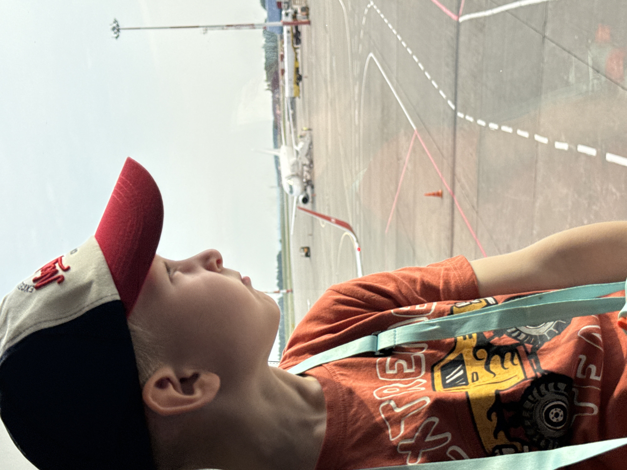 A young boy wearing a red, white, and blue baseball cap and an orange t-shirt with a graphic of a monster truck is looking out a window. The boy is positioned in the foreground, with his profile visible. The background shows an airport tarmac. Several airplanes are parked on the tarmac, including a white passenger jet with stairs extended to its door. A yellow and white refueling truck is also visible. The sky is overcast. The scene appears to be from an airport in or near Bol'shie Kabany, Russia.