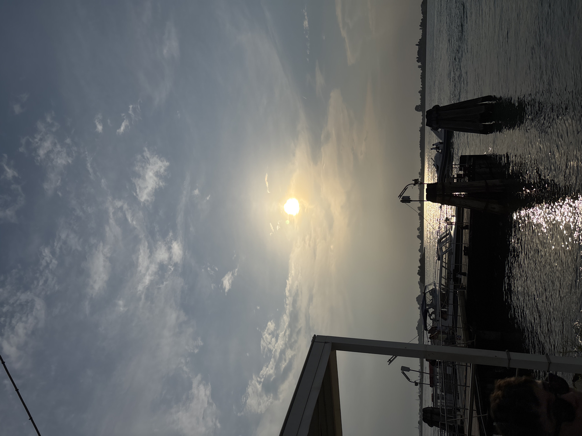 This photograph captures a scene in Punta Sabbioni, Italy, depicting a sun-drenched sky over water with various boats docked at a pier. The sun is a bright orb, positioned amidst scattered clouds in the sky, and its reflection creates a shimmering effect on the water's surface. Several boats are visible alongside the pier, their outlines blurred due to the bright light. Pilings stand in the water, supporting the dock structure. In the foreground, parts of a building frame and what appears to be the back of someone's head are partially visible, suggesting the photographer is under some structure and near people. The overall scene conveys a hazy, bright day at the waterfront.