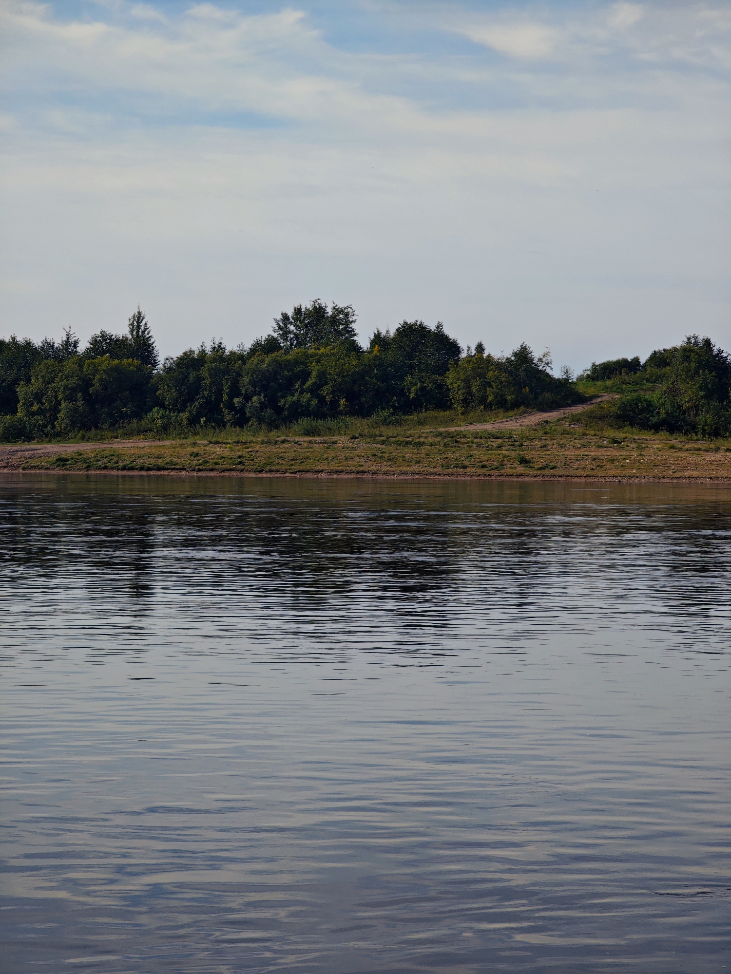 The image depicts a wide river with calm water reflecting the sky and the opposite bank. The far bank is lined with a dense growth of green trees and shrubs, with a dirt path winding up a slight incline. The sky above is partly cloudy with patches of blue. The scene appears to be a natural landscape, likely a riverfront area. This specific location is the Vyazma River in the city of Yartsevo, Smolensk Oblast, Russia, known for its scenic riverbanks and natural beauty.