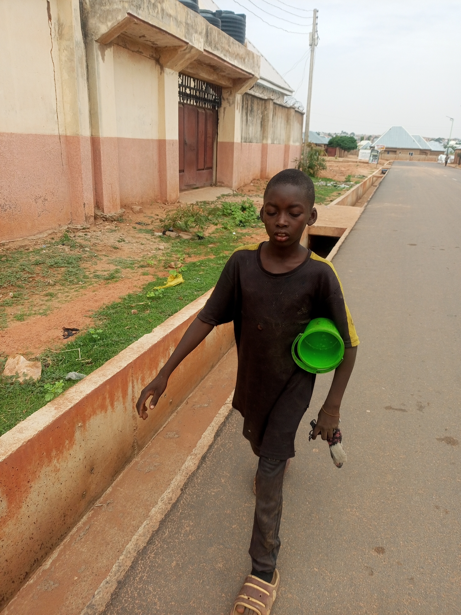 A young, thin Black boy walks along an asphalt street in Tempure, Nigeria. He carries a green bucket in his left arm and holds a small, sock-like object in his right hand. The boy wears a dark t-shirt with yellow shoulder stripes, dark pants, and brown sandals. He appears to be looking down.
To the left, a low concrete wall separates the street from a dirt and grassy area next to a two-story building. The building is light-colored with brown trim. A gated doorway is visible on the upper level. In the distance, other buildings and houses are seen under a cloudy sky. The street is mostly empty.