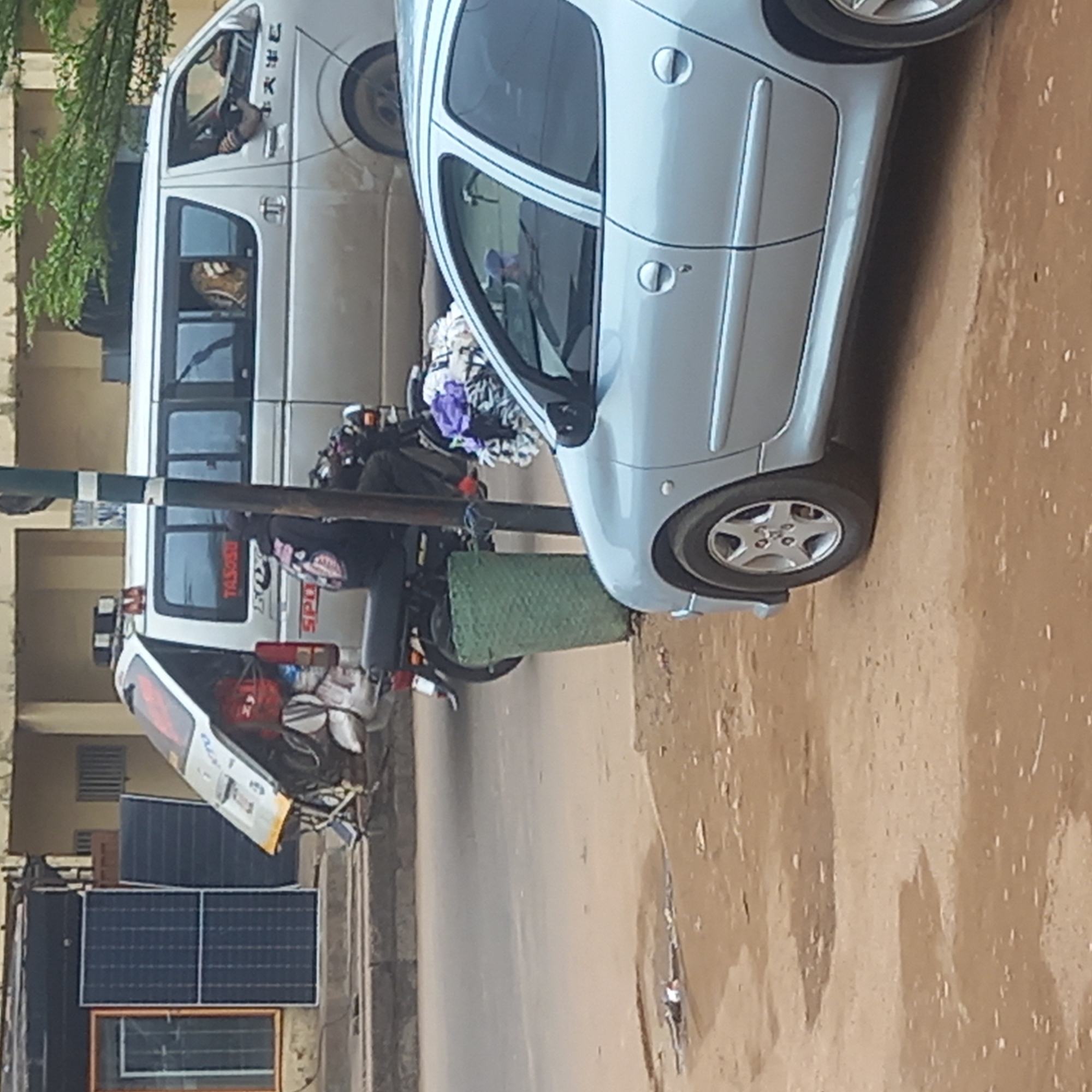 The image shows a street scene in Dadi Riba, Nigeria. A light blue compact car is parked on the right side of the road. A three-wheeled motorcycle with a white truck bed is parked to the left of the car, heavily loaded with goods. Another white van is parked behind the motorcycle. The street appears to be partially paved with a reddish-brown surface. Solar panels are visible on a building to the left of the motorcycle.