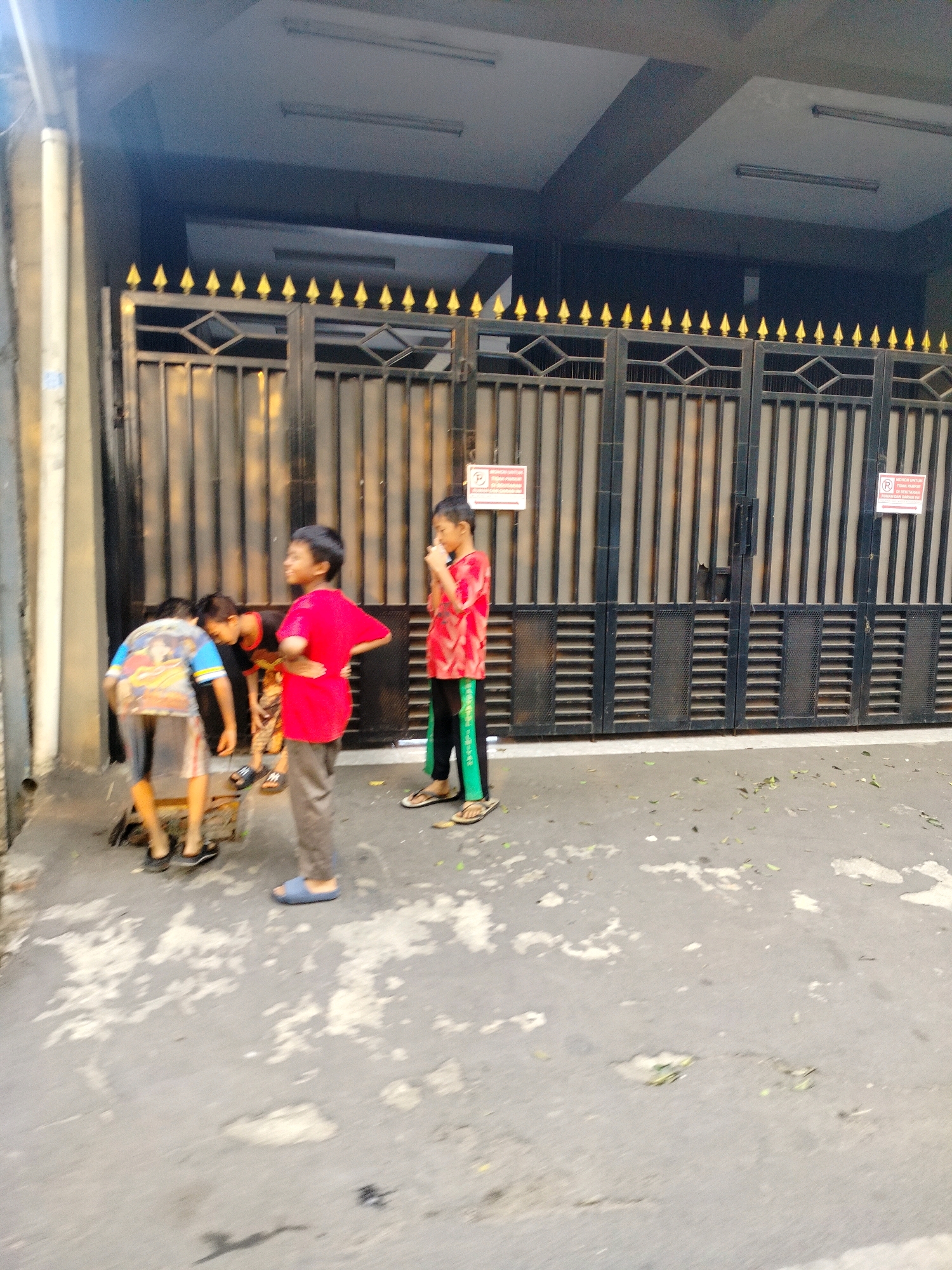 Four young boys are standing in front of a black metal gate with gold pointed accents. One boy in a blue shirt with a graphic print is bent over. Next to him, a boy in a red shirt is standing with his hands on his hips, looking to the left. To the right of him, another boy in a red shirt and black pants is looking down. The fourth boy, wearing a red and white patterned shirt and green pants, stands to the far right. The scene takes place on an outdoor concrete surface, likely a sidewalk or street, in front of a building in East Jakarta, Indonesia. The gate appears to be for a residential or commercial property. Two signs are visible on the gate, one on the left and one on the right, with text that is not legible in the image. The lighting suggests it is daytime, possibly late afternoon due to the shadows.