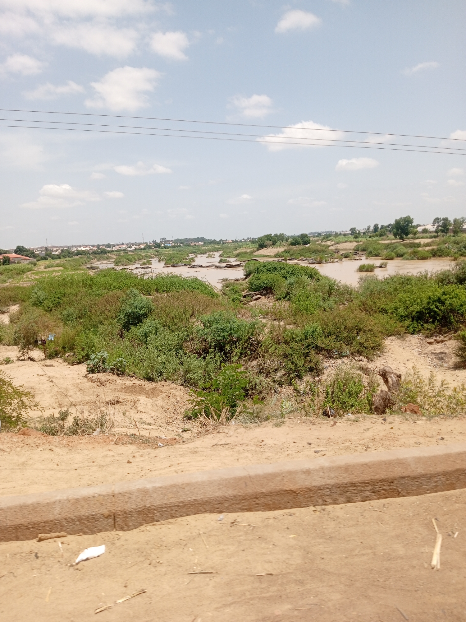 The image depicts a landscape in an unspecified city, in an unknown country. A muddy river runs through the scene, with sections obscured by green vegetation and scattered, dark, rock-like structures. The riverbanks are characterized by sandy and shrubby terrain. In the background, there's a settlement with buildings and sparse trees under a cloudy sky. Overhead, a pair of parallel power lines stretches across the top portion of the image. In the foreground, a sandy area is bordered by a concrete curb or low wall, with scattered debris including a small piece of white trash.