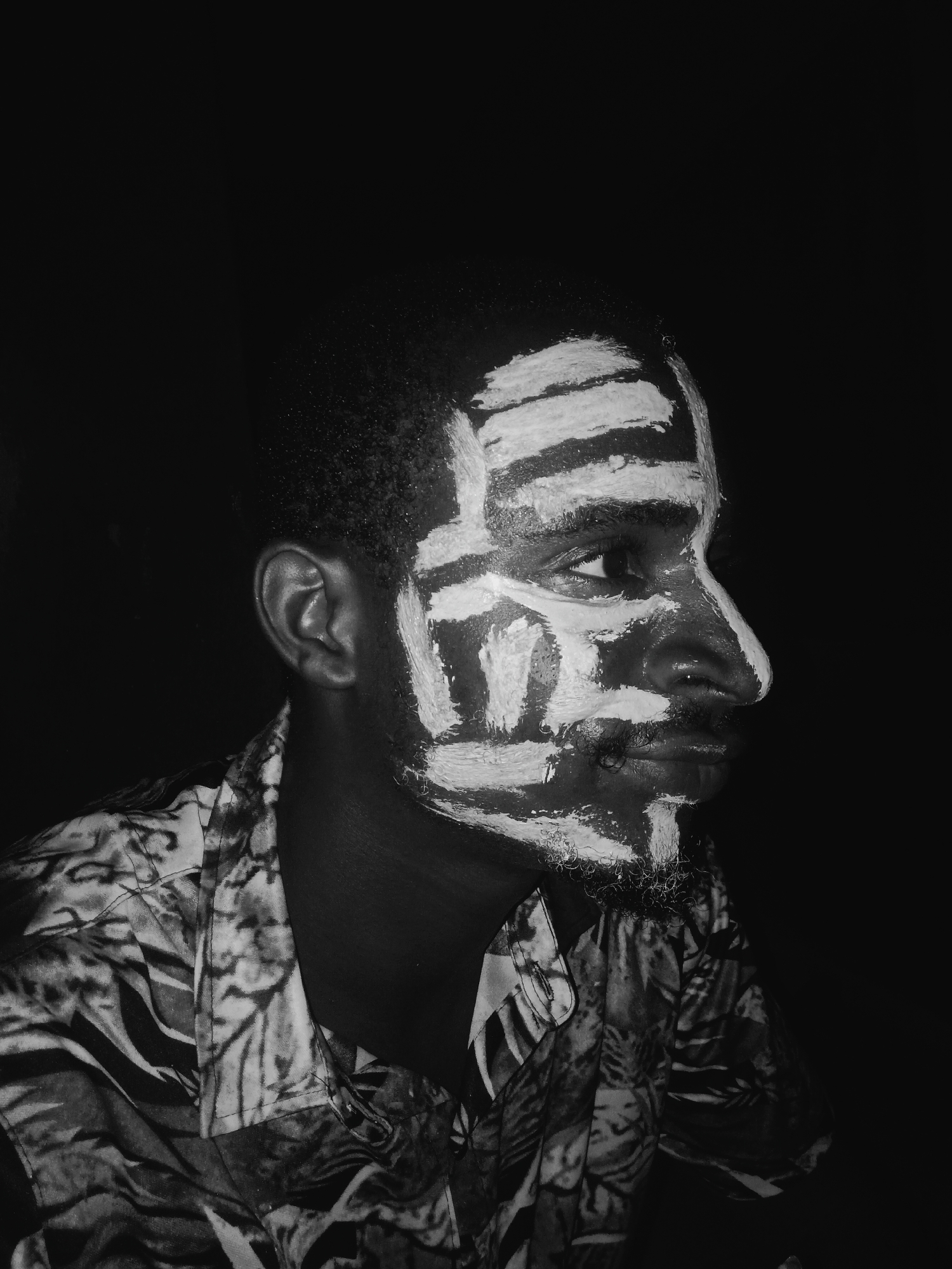A black and white portrait shows a dark-skinned man, possibly in Onitsha, Nigeria, given the prompt's instruction, with white paint covering sections of his face in a pattern of horizontal lines across his forehead and vertical lines on the rest of his face. He faces right, his expression contemplative. He is wearing a patterned shirt. The background is dark.