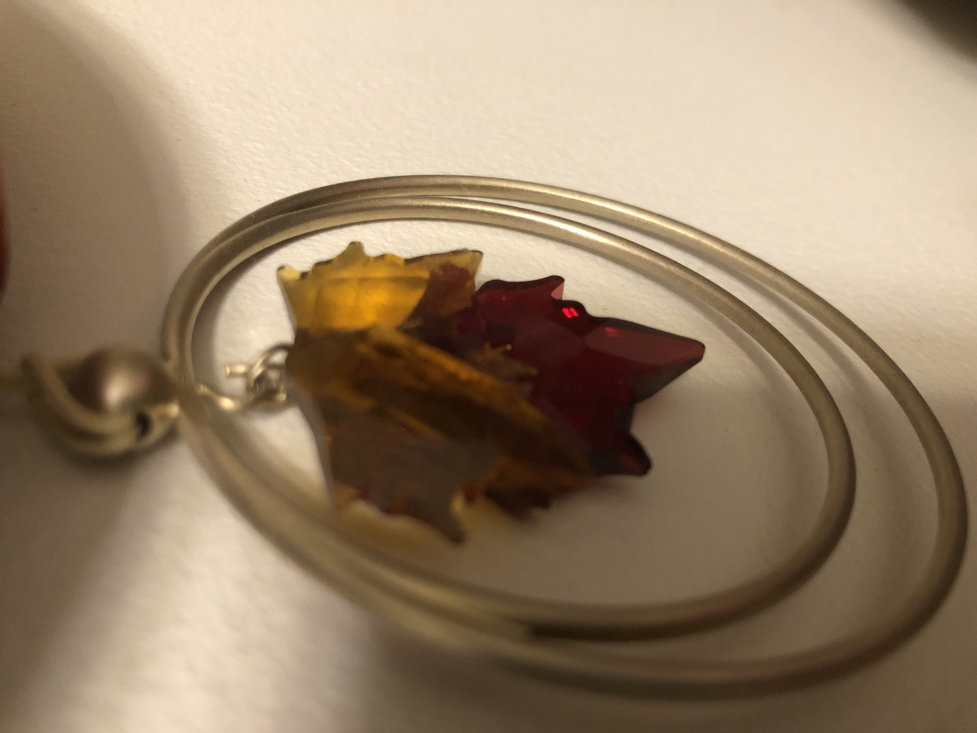 A close-up shot captures a pair of ornate gold-colored earrings. Each earring features a pendant in the shape of a maple leaf, crafted from multifaceted, translucent red and yellow resin. The leaves are suspended within a circular frame, also made of gold-colored metal, creating a sense of depth and elegance. The background is a plain, lightly textured wall, out of focus, which serves to highlight the intricate details of the earrings. The lighting is soft and diffused, casting subtle shadows that accentuate the reflective surfaces of the jewelry. The overall impression is one of refined craftsmanship and subtle autumnal beauty.