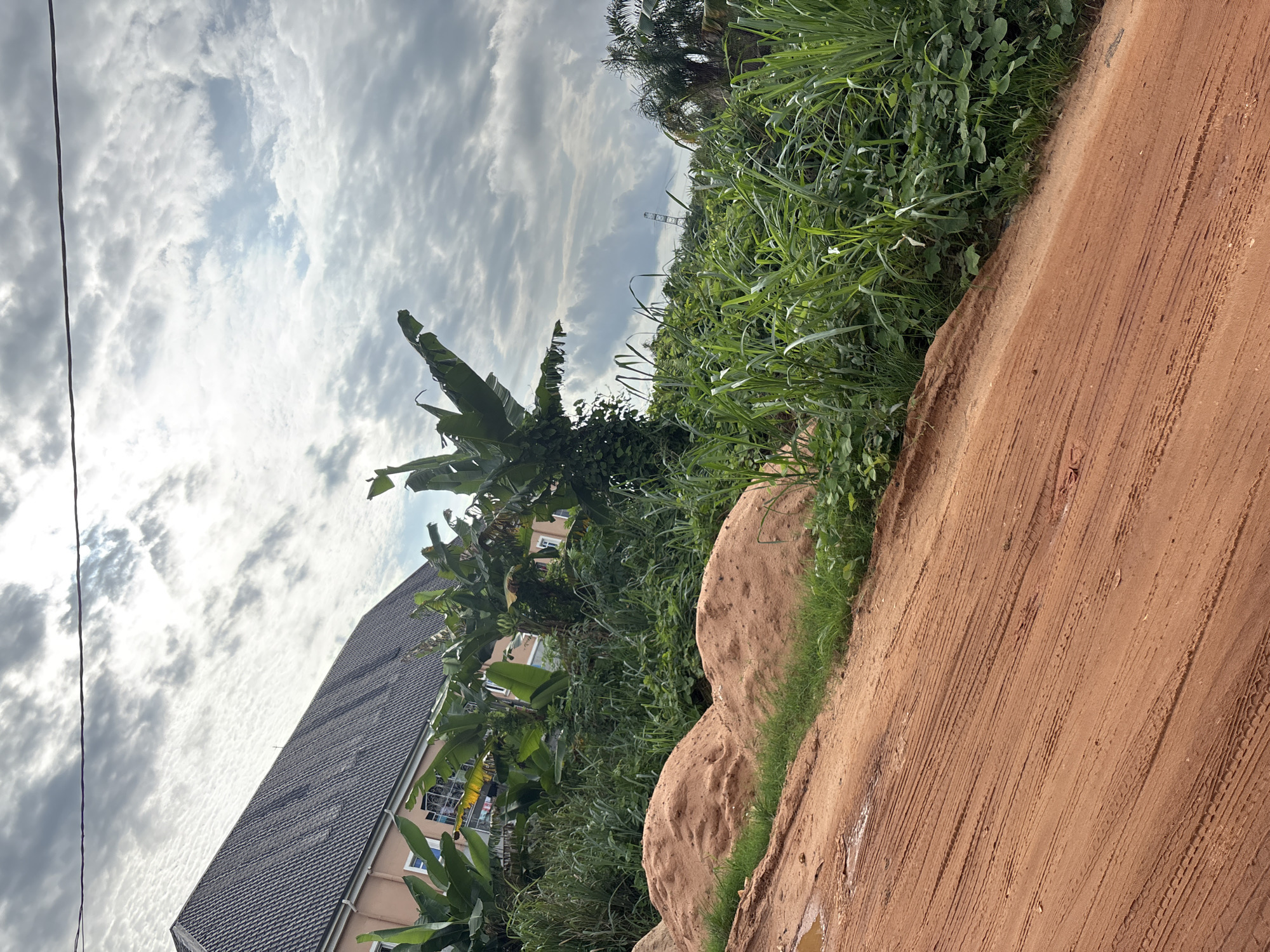 The image depicts a side view of a dusty red dirt road with tire tracks, leading uphill towards a landscape of lush green vegetation. In the background, a large building with a dark tiled roof and pale orange walls is visible, partially obscured by banana trees and other dense foliage. The sky above is cloudy, with patches of blue peeking through. The overall scene suggests a somewhat undeveloped area, possibly on the outskirts of the city of Obosi, Nigeria, where construction or landscaping activities might be taking place, indicated by the piles of sand near the road. There are no people or animals present in the image.