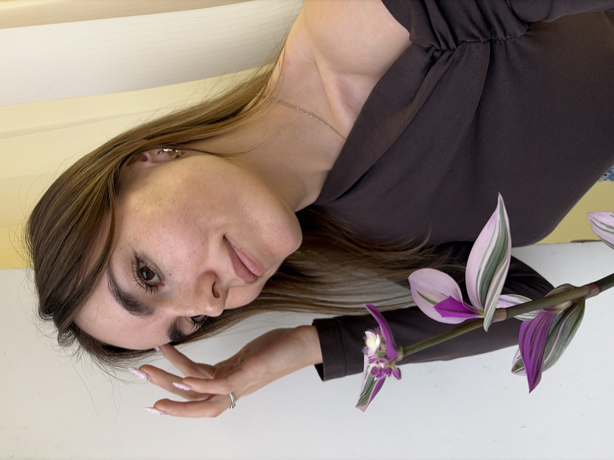A woman with long brown hair is holding a Tradescantia plant with purple, white, and green striped leaves and purple flowers. She is wearing a brown long-sleeved shirt. The background is a white wall or surface. There is no indication of Izhevsk, Russia.