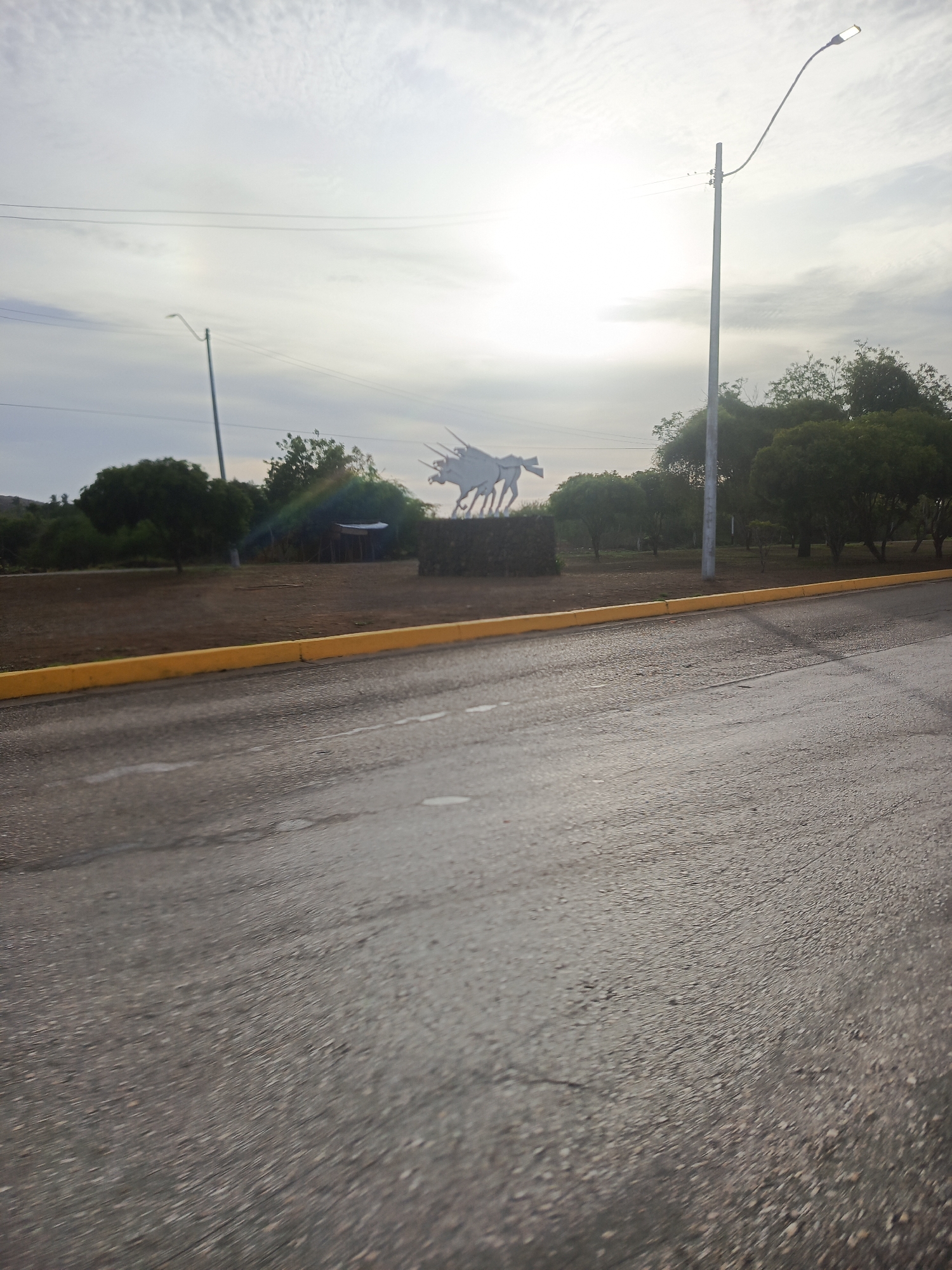The image depicts an outdoor scene near the Monumento a la Batalla de Matasiete in La Asunción, Nueva Esparta, Venezuela, likely captured from a moving vehicle. The foreground is dominated by a grey asphalt road exhibiting varied textures, suggesting surface irregularities or remnants of moisture, bordered on the left by a yellow concrete curb. Beyond the curb, a strip of dry, light-brown earth with sparse vegetation leads to a cluster of green trees and bushes of varying sizes and densities.
In the midground, a tall, slender utility pole supports power lines and a street light with a curved arm extending to the upper right. Another, smaller street light is visible further to the left. Central to the scene, a large, light-colored metallic sculpture of multiple horses in dynamic motion is positioned on a low, dark base. The horses appear to be galloping or charging, with elongated, possibly spear-like elements extending from some forms. Behind and to the left of the sculpture, partially obscured by trees, is a rustic, low-lying structure with a light-colored roof and dark support poles.
The sky in the background is bright and predominantly overcast with scattered clouds, creating a diffuse light source. A prominent multicolored lens flare is visible in the upper mid-right portion of the image, indicating the presence of a strong light source, likely the sun, near the frame's edge. Distant hills or elevated terrain are faintly visible on the far left horizon. No individuals or active animals are present in the frame.
