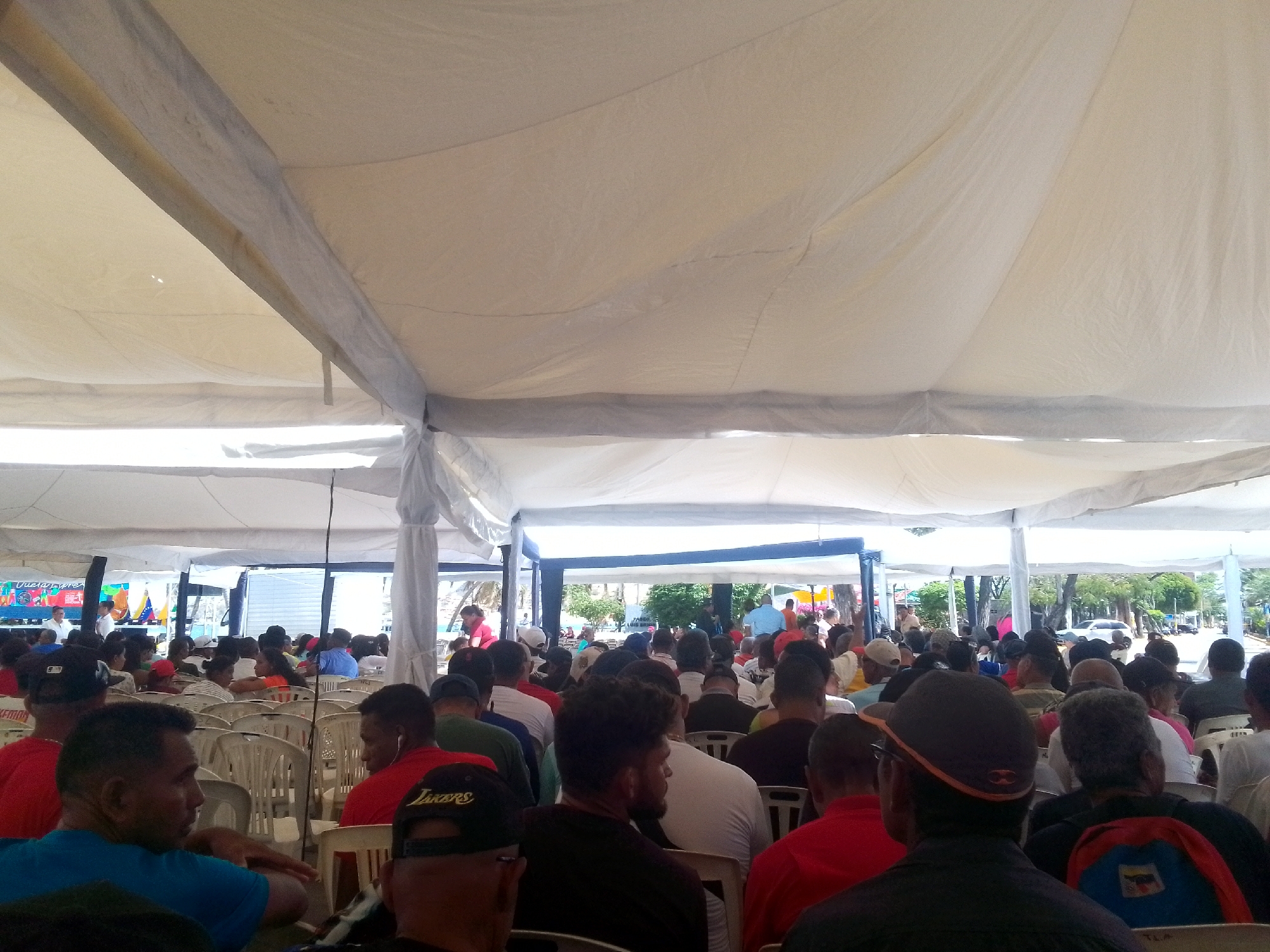 A gathering of people is taking place under a large white tent-like structure. The scene appears to be set in an outdoor environment, potentially a public square or park in Juan Griego, Venezuela. Numerous individuals are seated in rows of white plastic chairs, facing a direction obscured from the viewer's perspective. A variety of clothing can be observed on the people in attendance, including hats, t-shirts, and backpacks. The event is held during the day and the sky is clear.