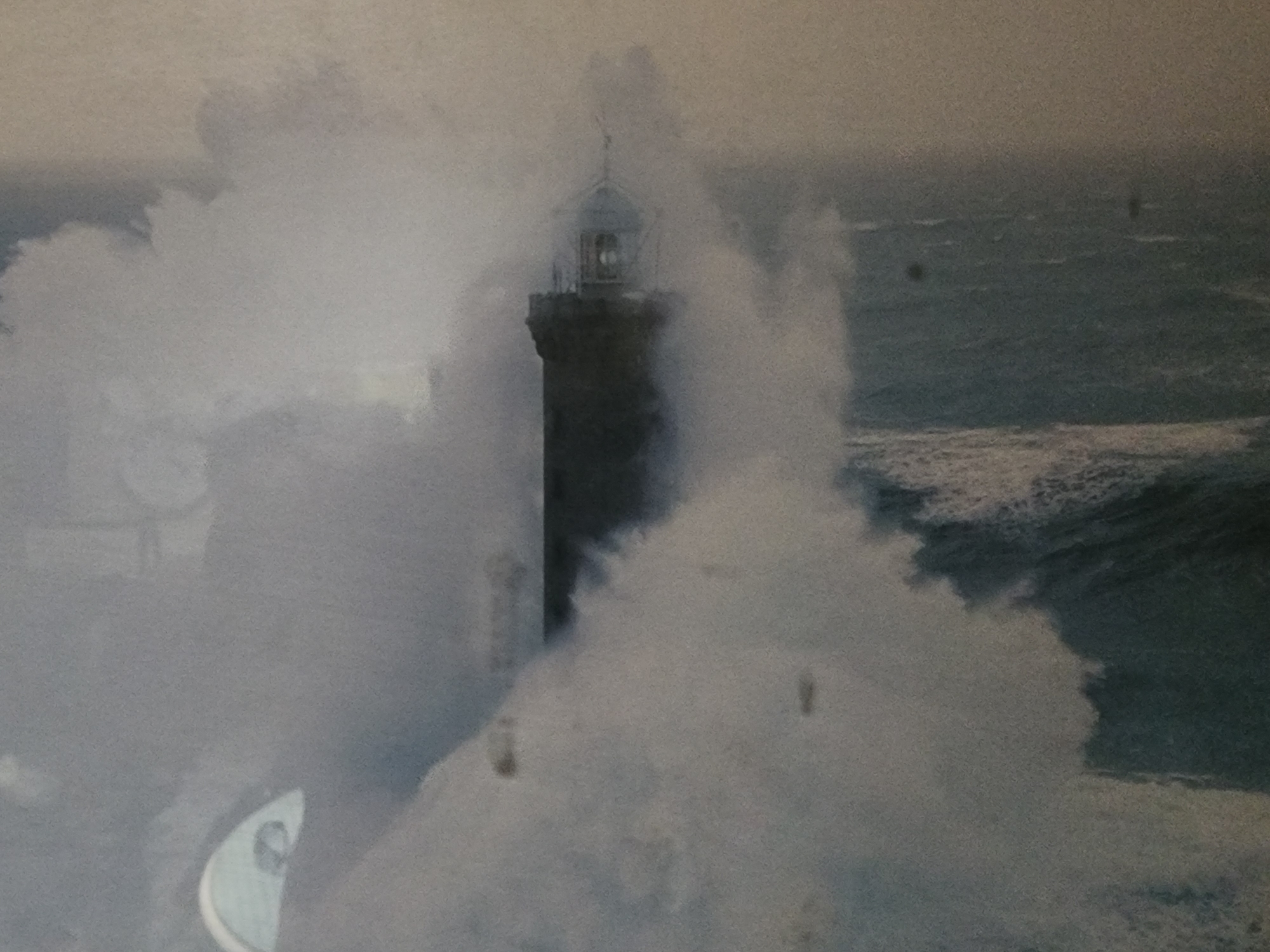A lighthouse stands tall in the center of the frame, buffeted by a powerful wave that envelops its base and obscures much of its structure in a dense spray of white water. The lighthouse appears to be constructed of dark stone and has a lantern room at its peak, visible through the mist. The sea is rough, with large waves crashing around the lighthouse. The overall atmosphere is dramatic and turbulent, suggesting a storm or high winds. The image is desaturated, giving it a muted, monochromatic appearance. The setting is likely a coastal area.