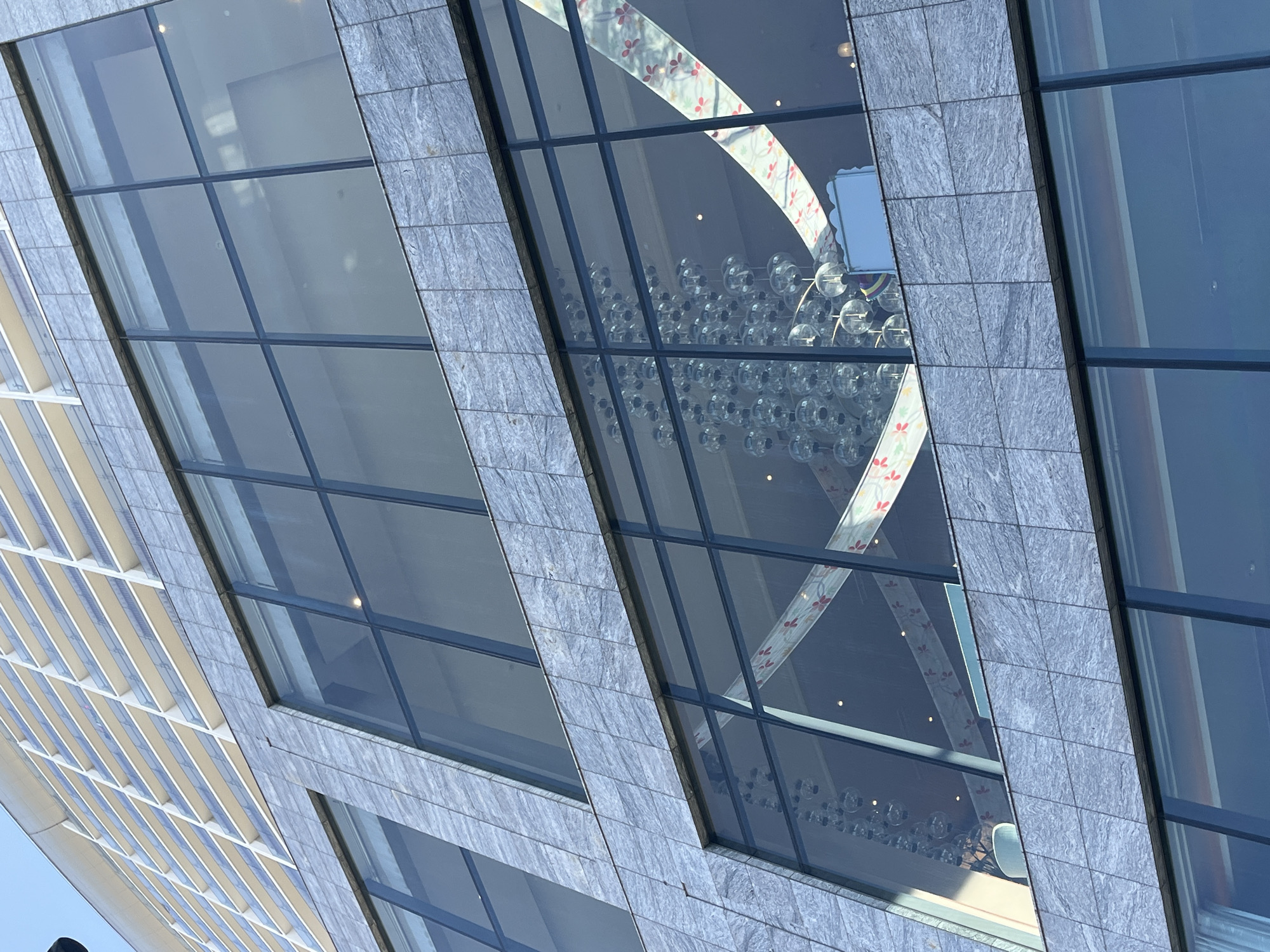The image presents a close-up, diagonally oriented view of a modern building facade in Đà Nẵng, Vietnam. The building features large rectangular glass panels framed by sections of grey, textured stone cladding.
Through the central windows, the interior of the building is visible, showcasing a large, ornate chandelier composed of numerous clear spherical glass lights. Above the chandelier, a decorative architectural element, possibly an archway or ceiling feature, is visible. This feature is white with a repeating pattern of small red and white flowers.
The glass panels also reflect a bright blue sky and the facade of another building to the left, which has a pattern of light-colored horizontal slats or louvers. The strong, clear lighting suggests it is daytime with good weather. There are no people visible, nor is there any discernible activity or text.