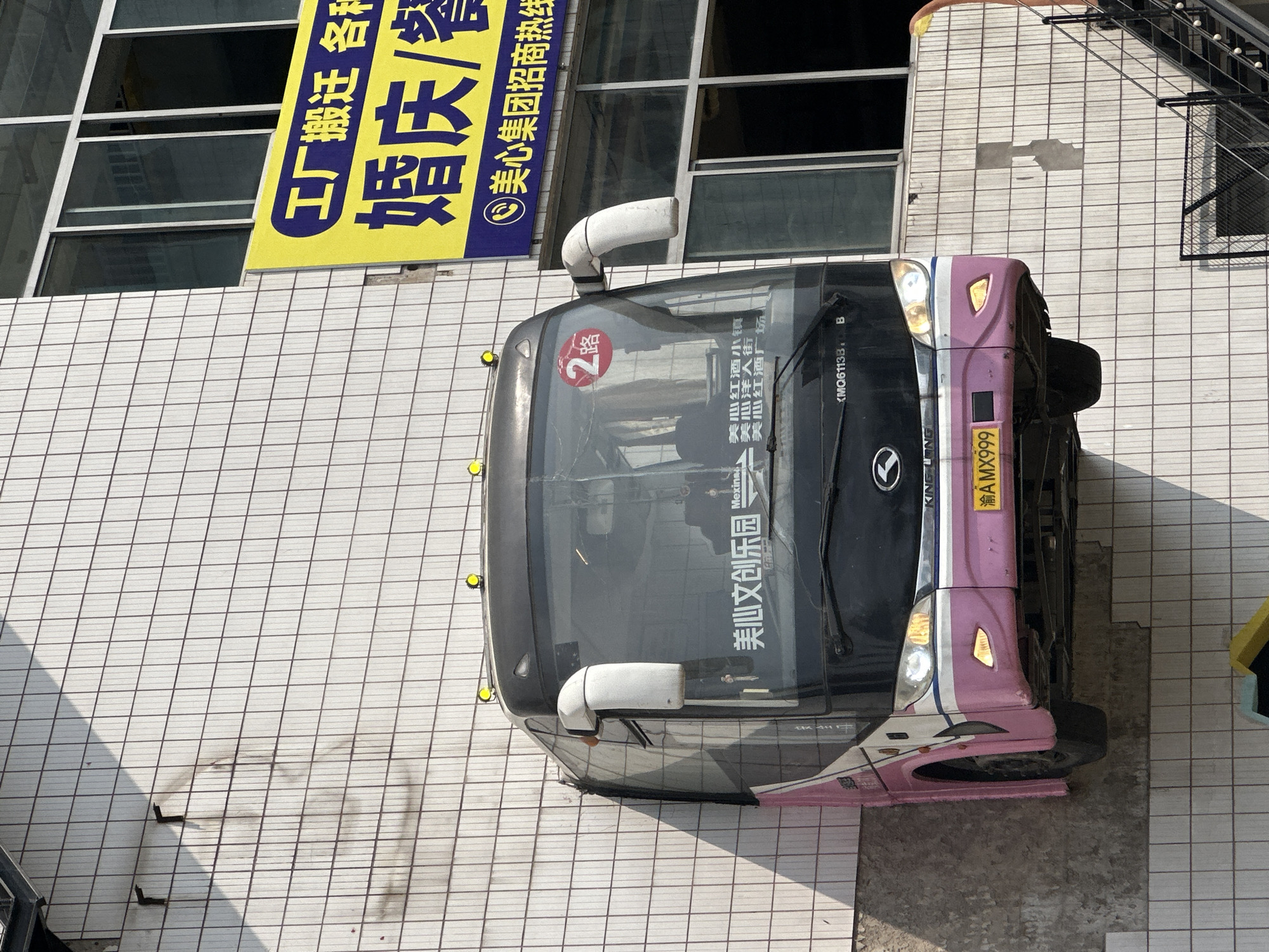 A bus is photographed outdoors, parked near the side of a building with signage. The bus has a pink and black exterior and the windshield displays "美心文创乐园" (Mexin Creative Park), "2路" (Route 2), and "美心红酒小镇 美心洋人街 美心红酒广场" (Mexin Wine Town, Mexin Foreigner Street, Mexin Wine Plaza) in Chinese characters. The license plate is "渝AMX999." The building beside the bus has a yellow sign with blue Chinese characters. A staircase is seen on the right side of the building. The location is in Nan'an District, China.